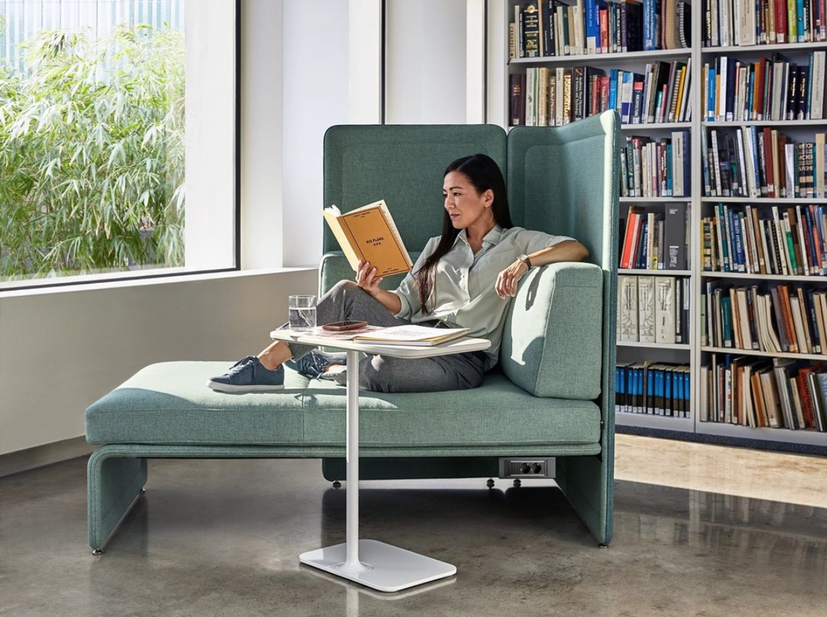 A woman sitting comfortably in a modern library or study area, reading a book. She is seated on a teal, high-backed booth with a small white table in front of her, which holds a glass of water and some opened books or notebooks. The background features a large window with greenery outside and a bookshelf filled with numerous books.