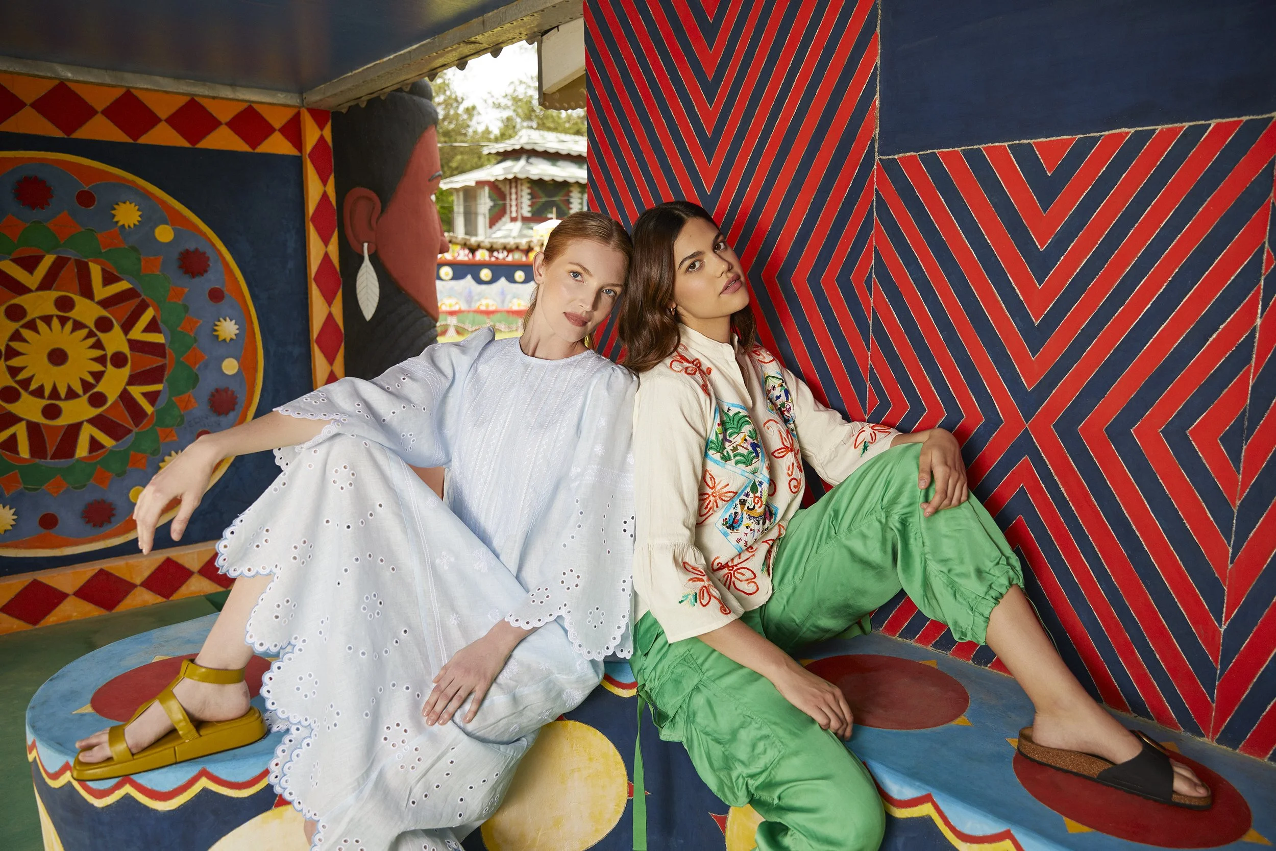 Two women sitting on colorful, patterned seats with vibrant murals behind them at an amusement park.