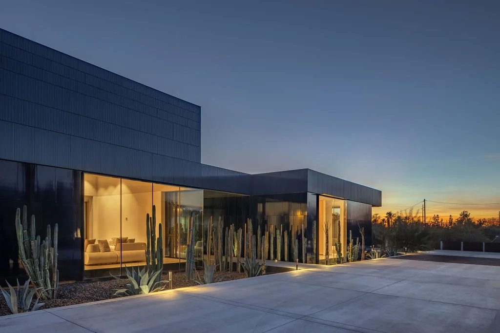 Modern house with black exterior walls, glass windows, and desert plants outside during sunset.