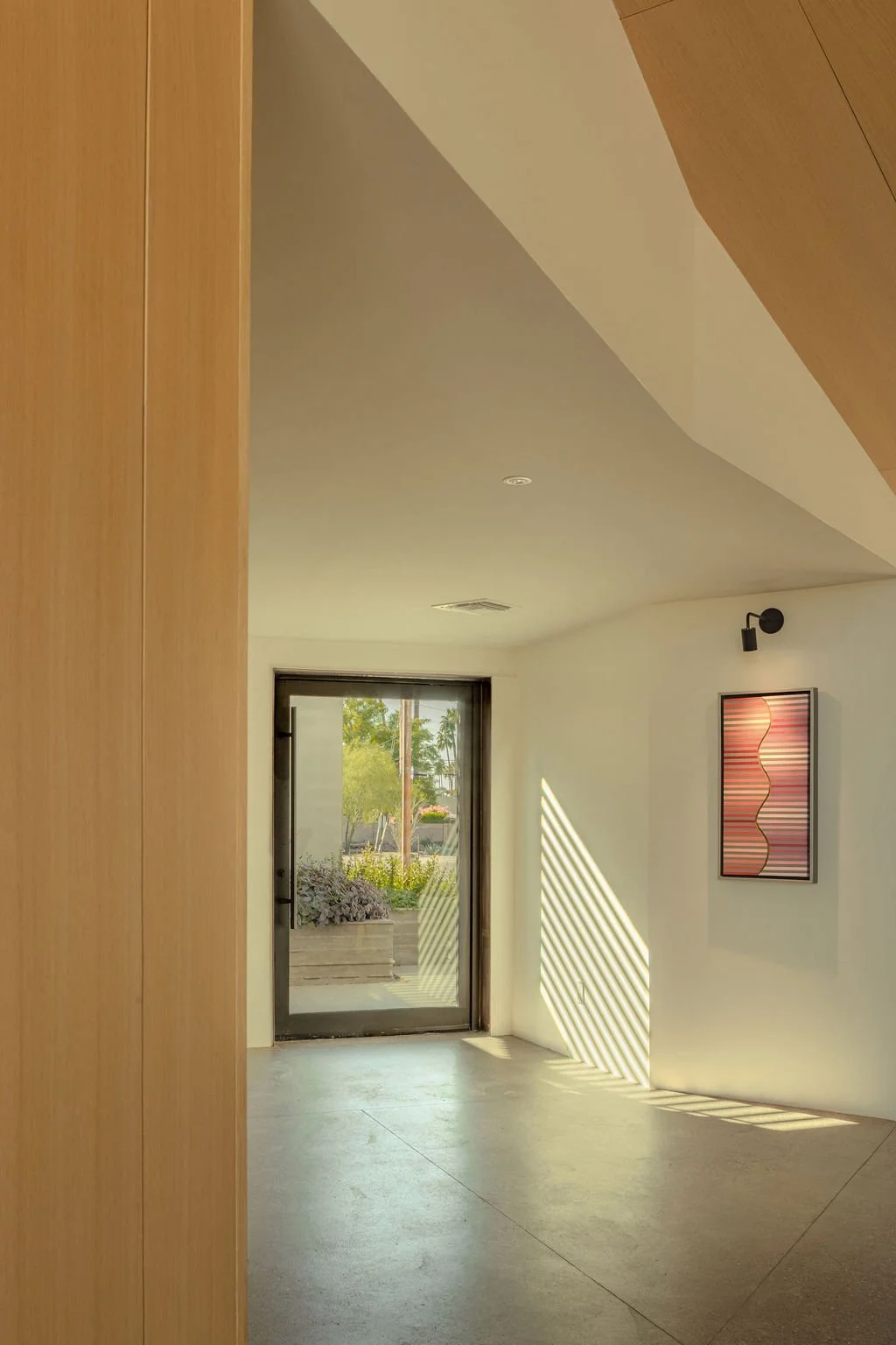 Sunlight coming through a glass door creates striped shadows on a white wall, with a framed abstract artwork hanging above. A black wall light is mounted near the artwork.