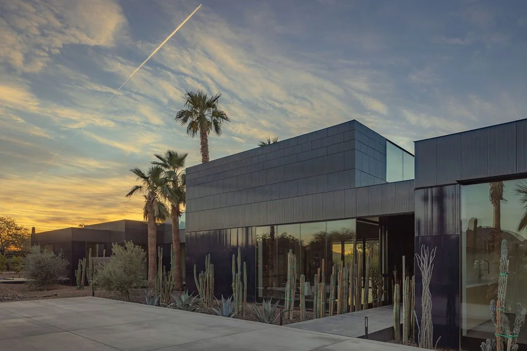 Modern building with dark exterior, palm trees, and cacti in a desert landscape at sunset.