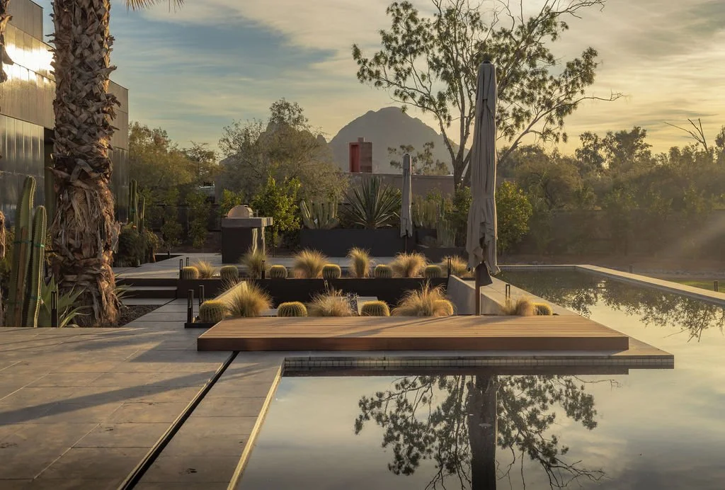 A modern outdoor pool area at sunset with cacti, trees, and mountains in the background, featuring a pool, wooden deck, sun umbrellas, and seating areas.