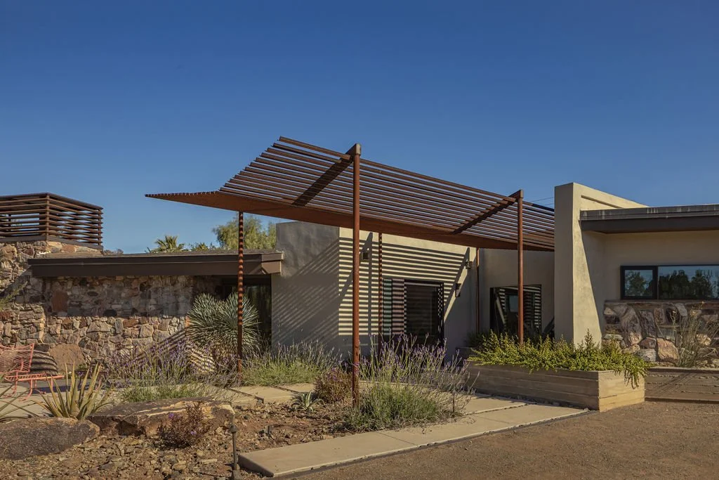 Modern house with stone and stucco exterior and a wooden pergola in a desert landscape with drought-tolerant plants.