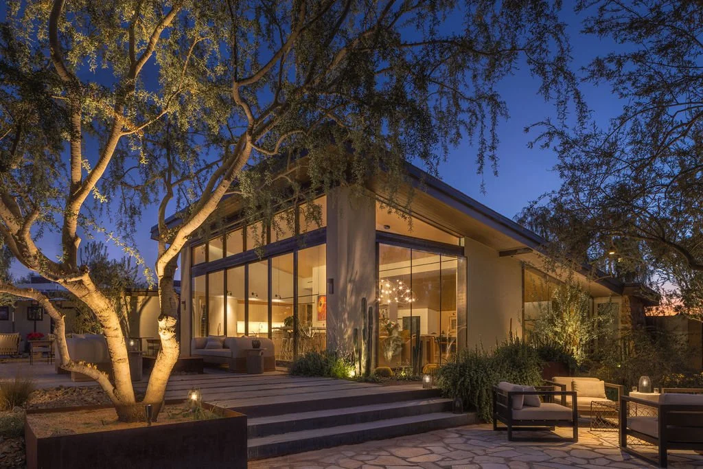 Modern house with large glass windows and door, illuminated interior, surrounded by trees and outdoor seating, during dusk