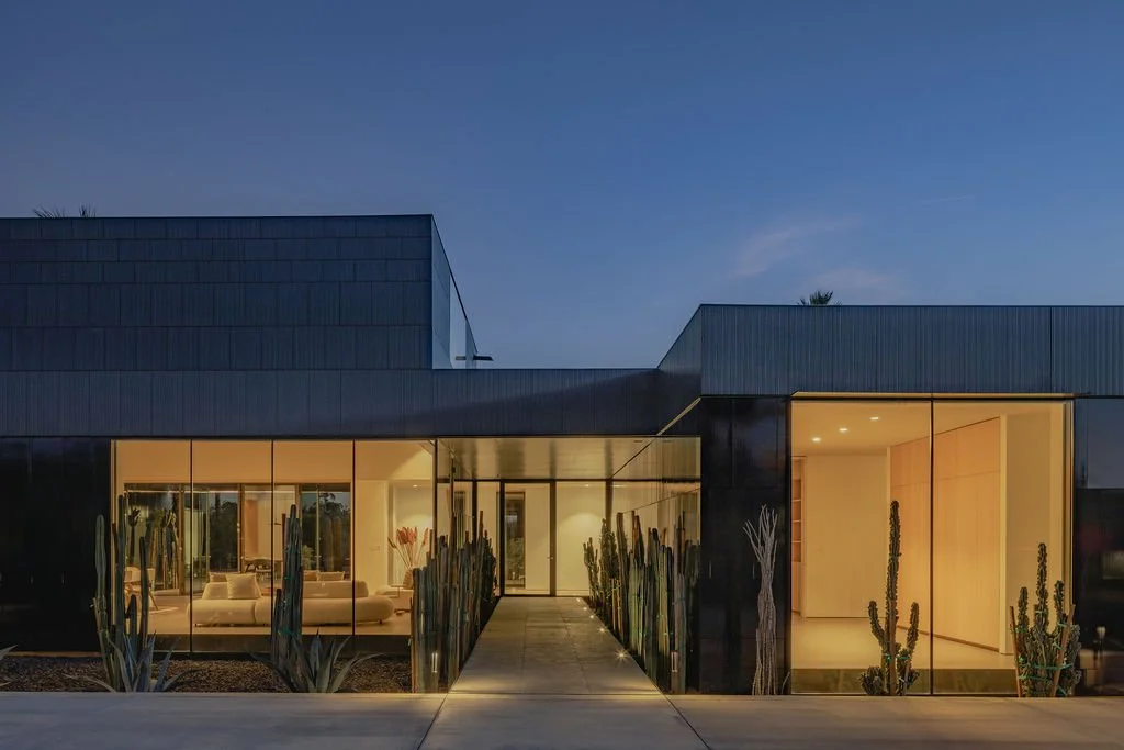 Modern house with large glass windows illuminated from inside, surrounded by desert plants and cacti, under a clear evening sky.
