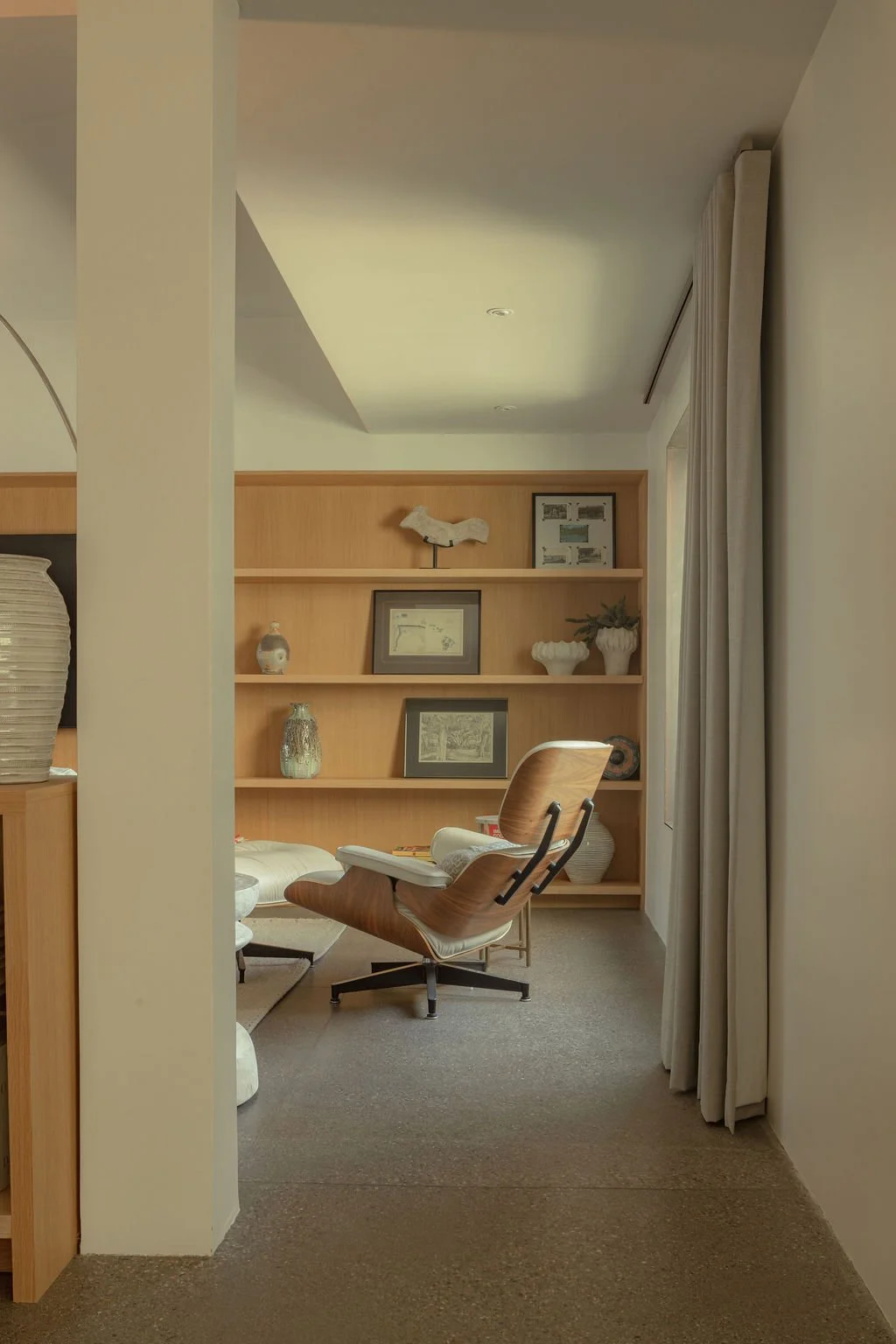 A cozy reading nook with wooden shelves, framed artwork, decorative vases, and a modern chair with a curved wooden backrest and white cushion, next to floor-length curtains.