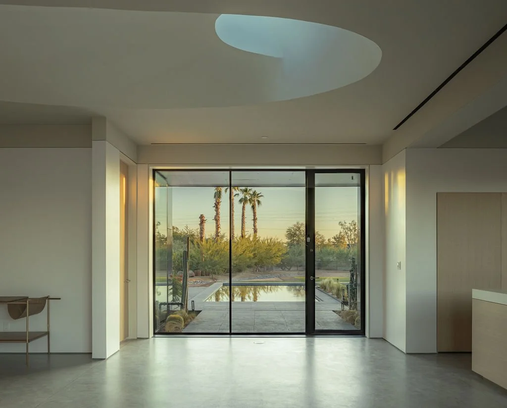Modern living room with large sliding glass doors leading to a backyard with a swimming pool, desert landscaping, and palm trees, in the late afternoon or early evening.