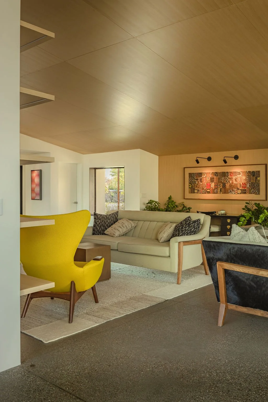 Living room with cream sofa, yellow armchair, black armchair, decorative pillows, large artwork on wall, green plants, ceiling lights, windows, and textured rug.