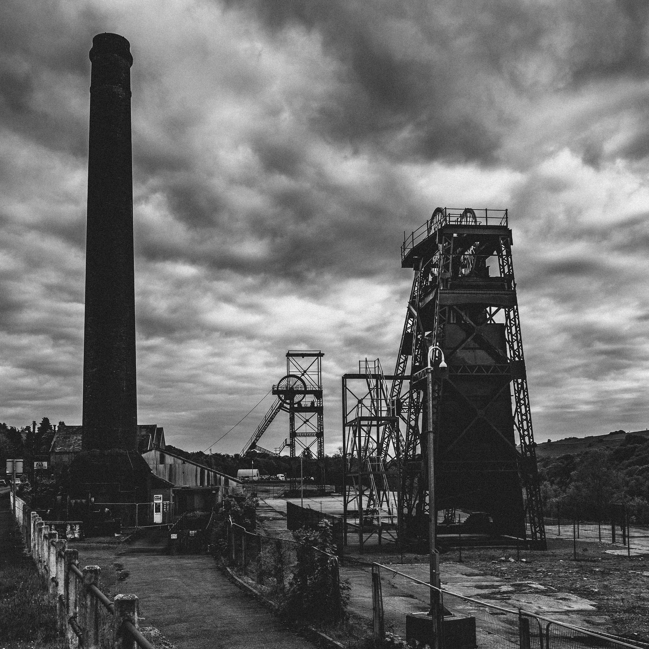 Wide-angle black and white photograph of Cefn Coed Colliery chimney with twin winding towers and cloudy sky.
