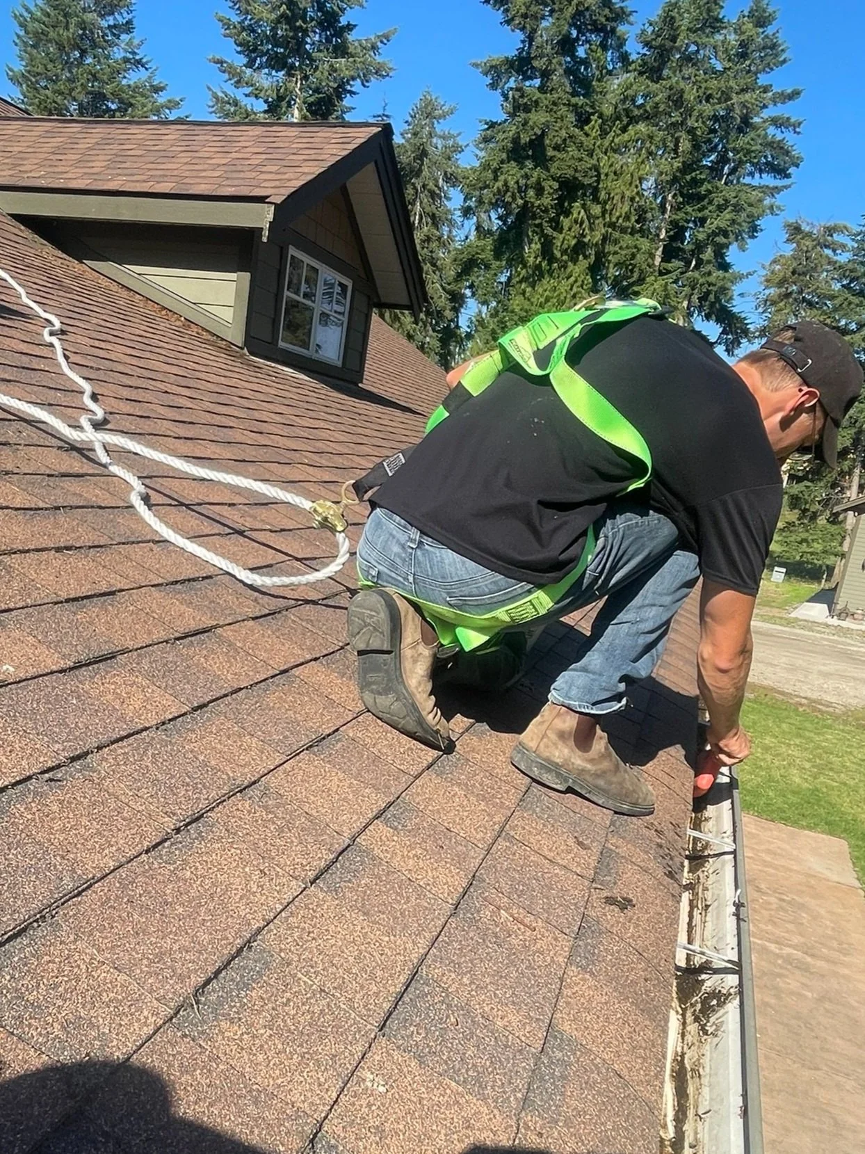 A man wearing a black shirt, jeans, and work boots, kneeling on a roof, working with a tool near the gutter, with a safety harness attached to a rope for fall protection.
