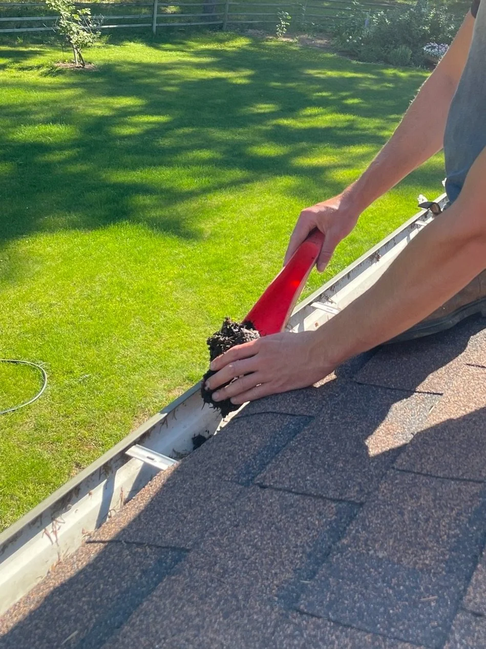 A person cleaning the gutter of a roof with a red scoop using a black scrub brush, with a green lawn and trees in the background.