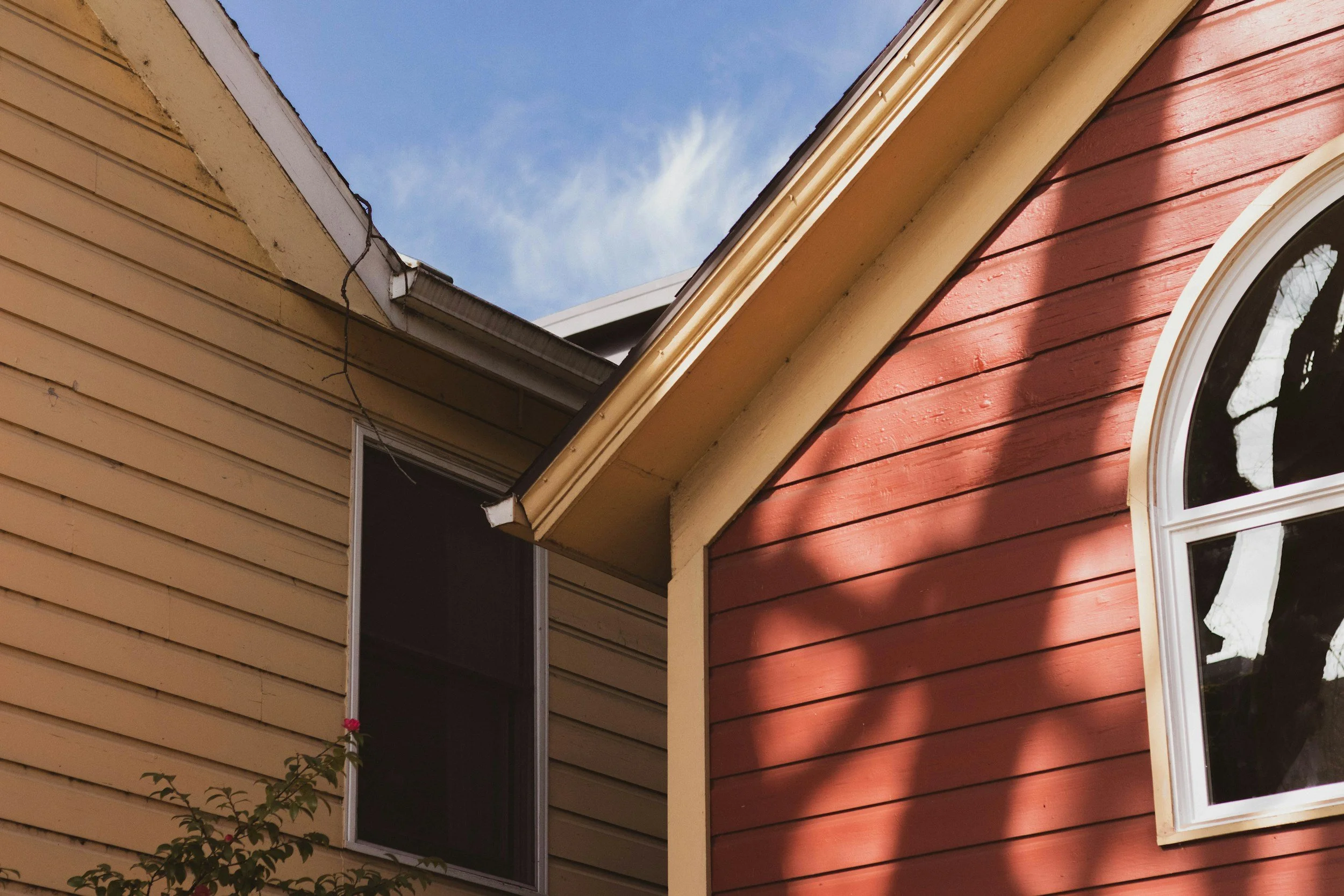 Close-up of two houses with different colored siding, one yellow and one red, showing part of the roofs and windows, with a tree's shadow on the red house and a blue sky in the background.