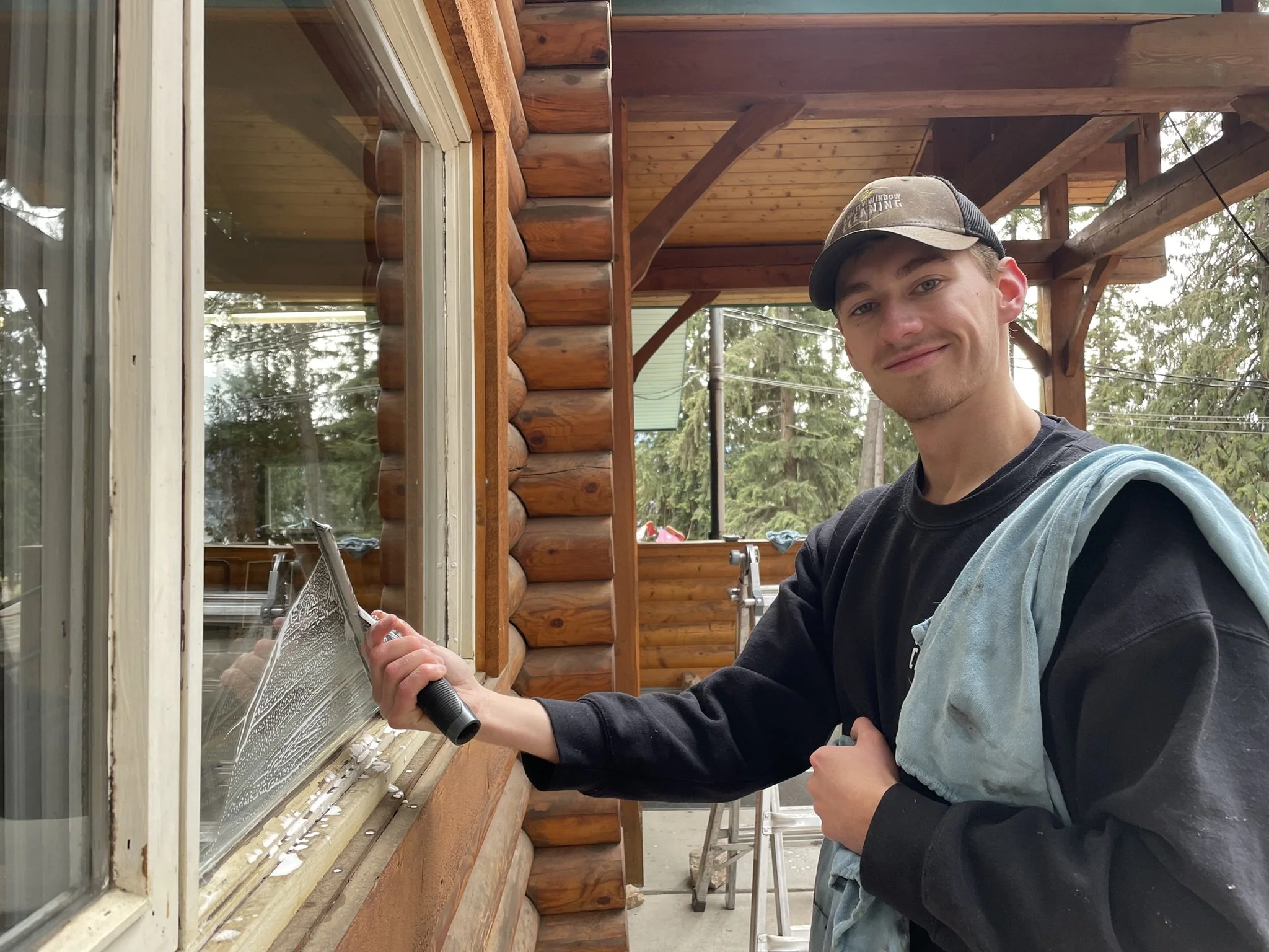 A young man smiling while cleaning a large window on a log cabin exterior during daytime, holding a cleaning tool, with a ladder and trees in the background.