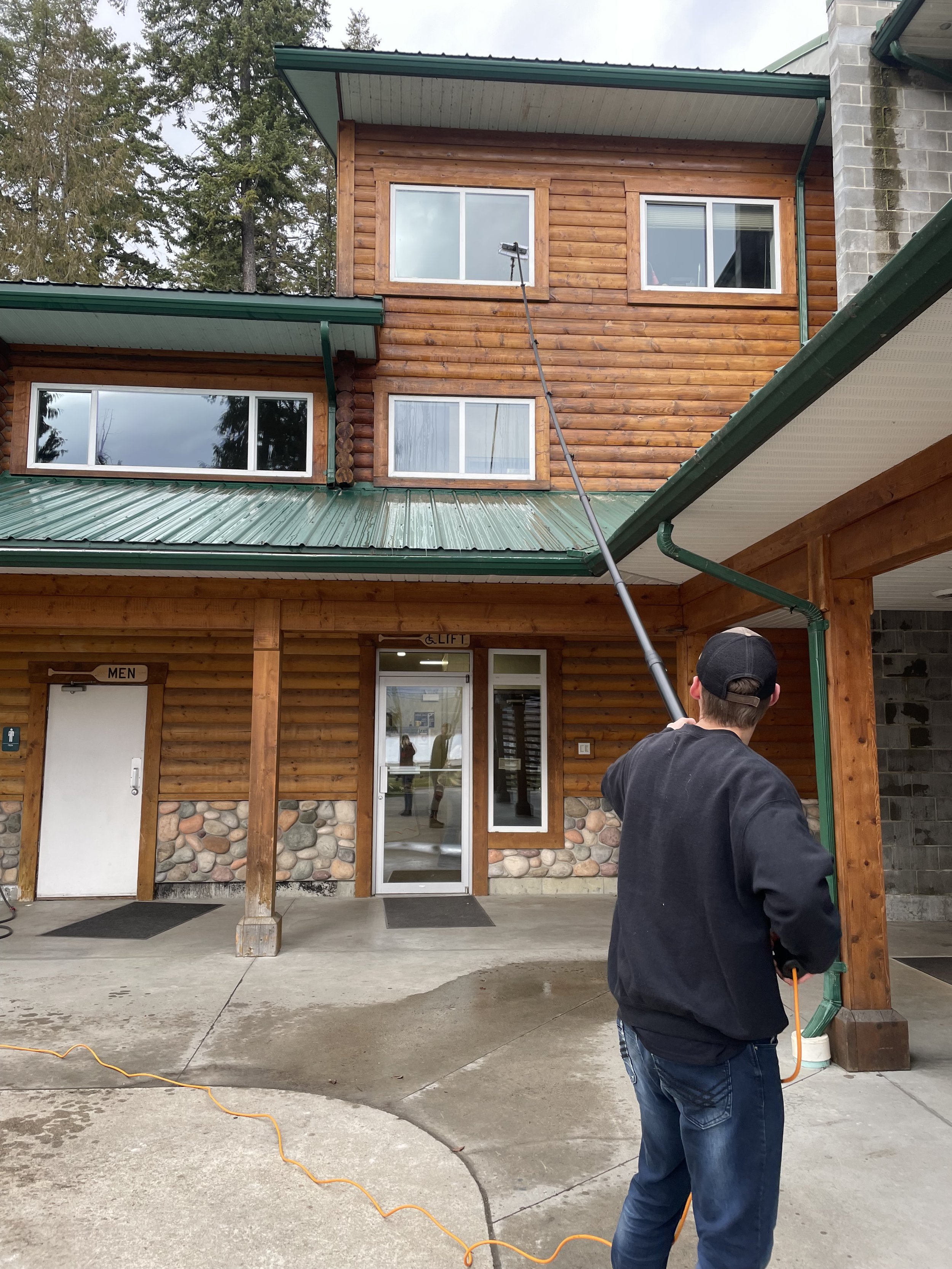 A person pressure washing the exterior wood siding of a building with multiple windows and a green metal roof.