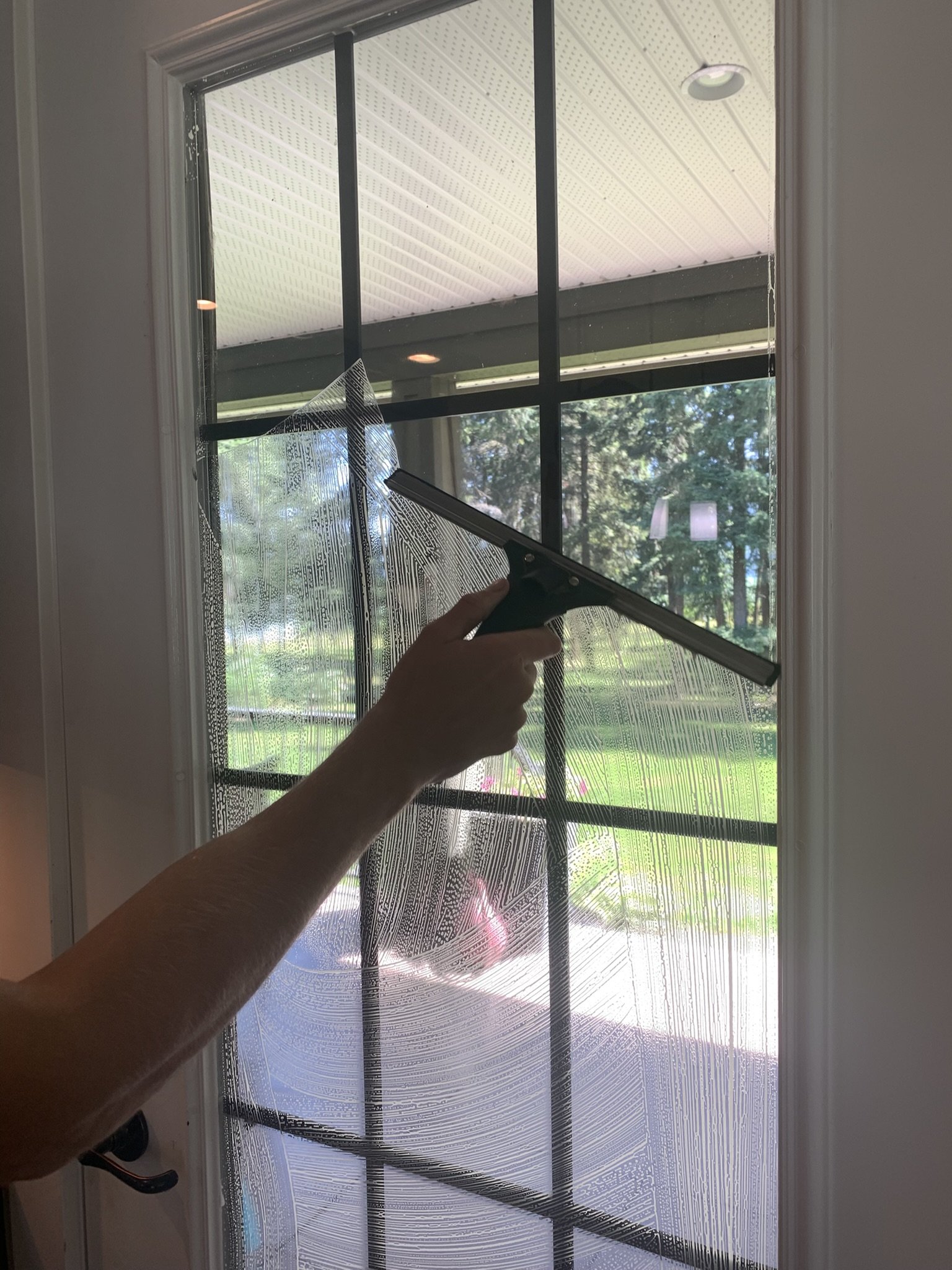 Person cleaning a window with a squeegee, creating soap streaks on the glass, with a view of green trees and a lawn outside.
