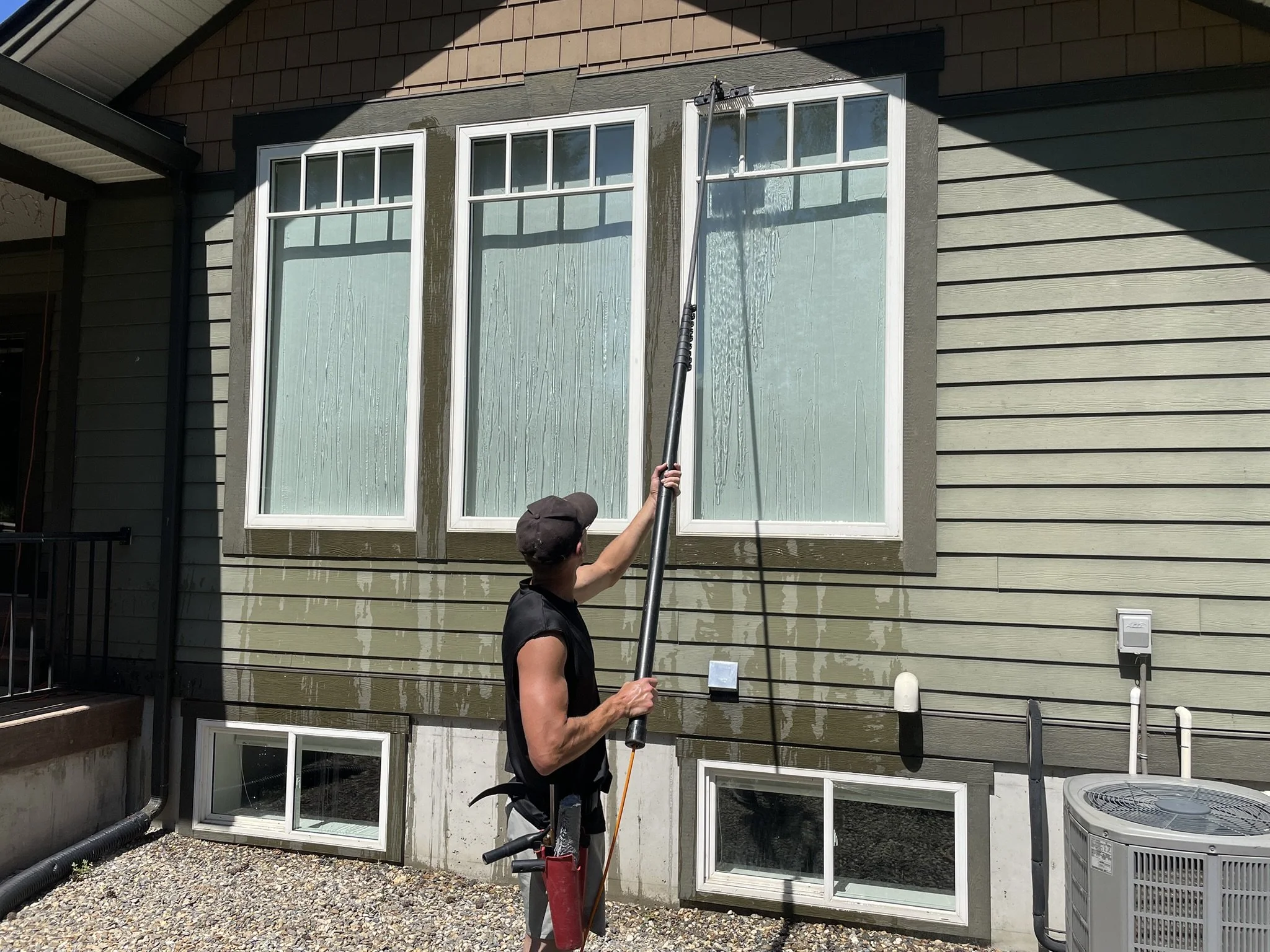 Person power washing the exterior of a house with green siding and large windows.