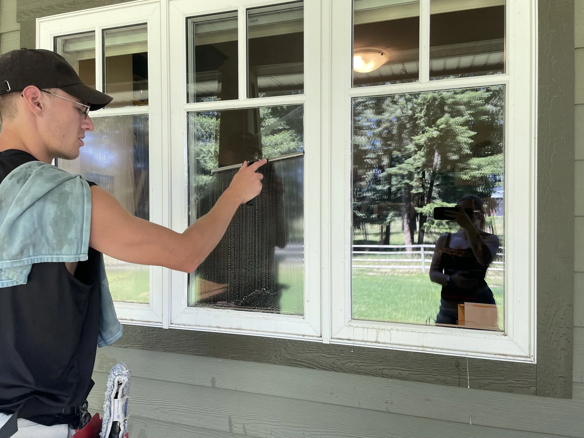 A person cleaning a window with a squeegee outside a house during daytime, with trees and a white fence reflected in the glass.
