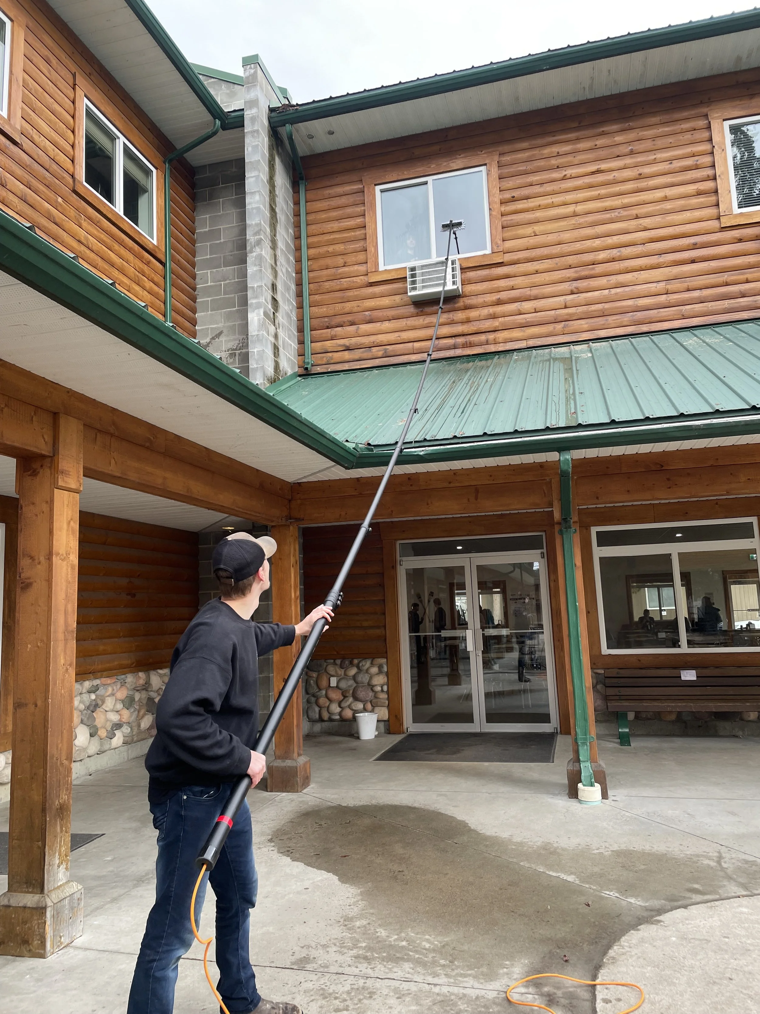 A person pressure washing the concrete patio area outside a wooden building with trees reflected in the glass doors.