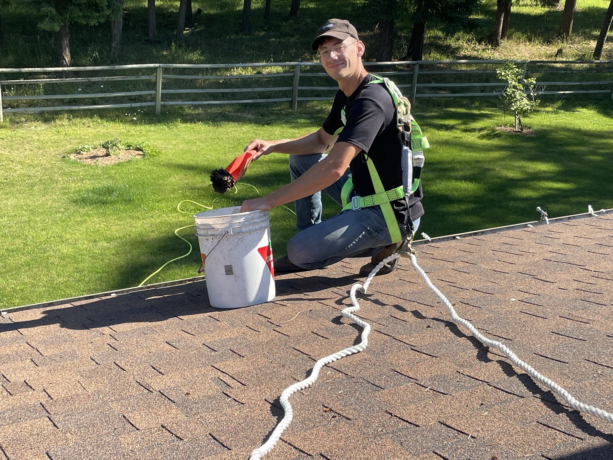 A man kneeling on a rooftop, wearing a harness, holding a bundle of wires or cables, and smiling at the camera. He is on a shingled roof with a bucket nearby and a green yard with a fence and trees in the background.