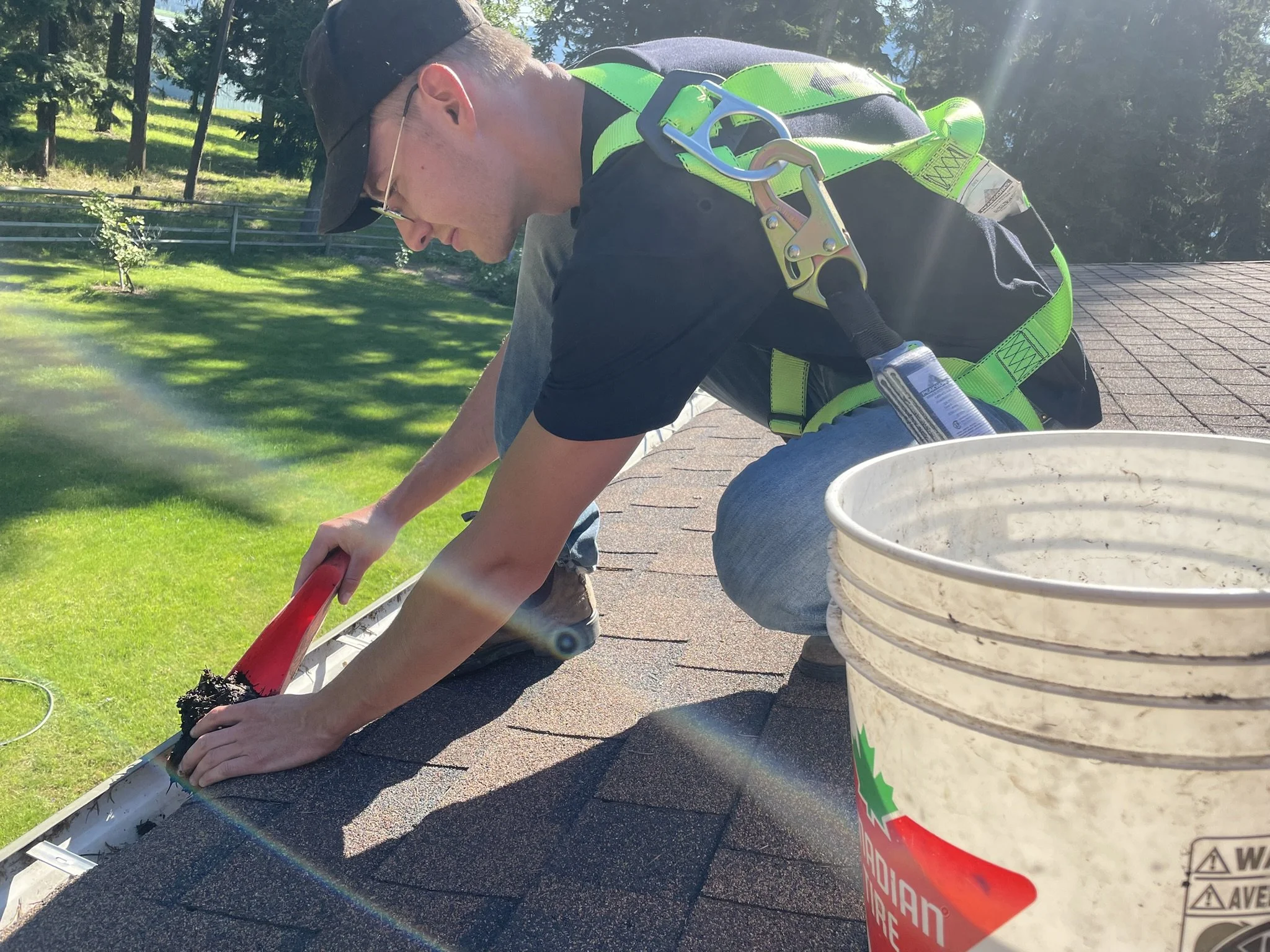 A person wearing a safety harness cleaning a gutter with a scrub brush and a red-handled tool on a roof during daytime, with a bucket nearby and a green yard in the background.