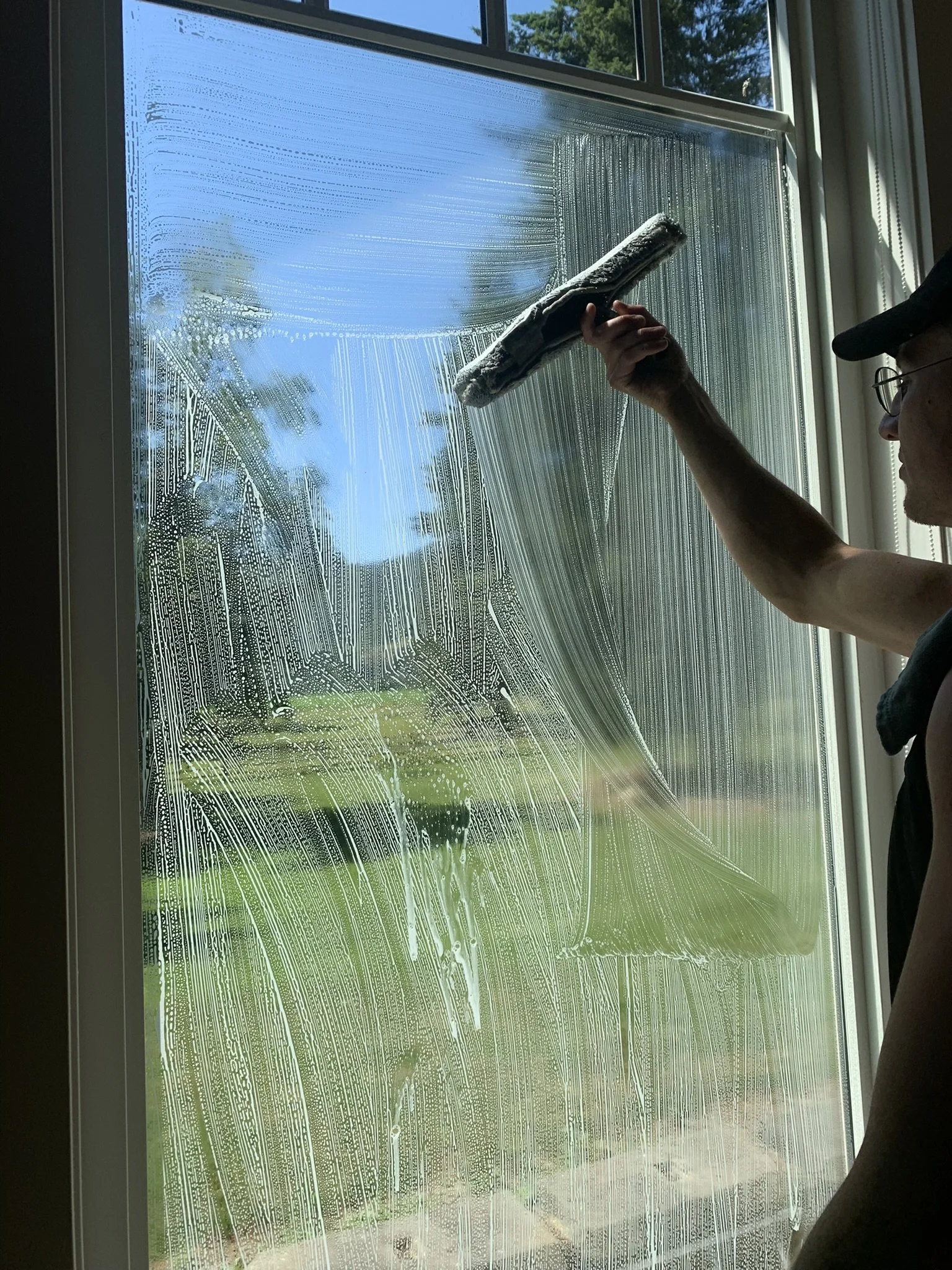 Person cleaning a window with a squeegee, creating soap suds and streaks, with a view of trees and blue sky outside.