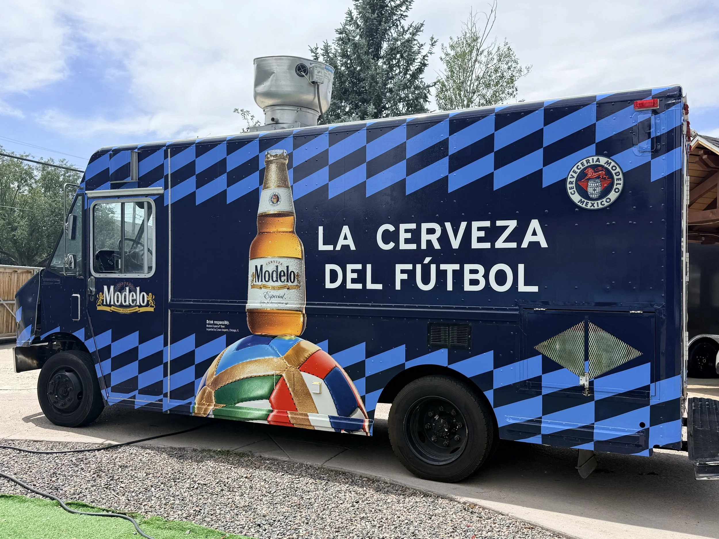 A food truck with blue geometric pattern and a beer advertisement, featuring a large beer bottle image, with a volleyball decorated with the Mexican flag colors in front, and green turf adjacent to gravel ground under a partly cloudy sky.