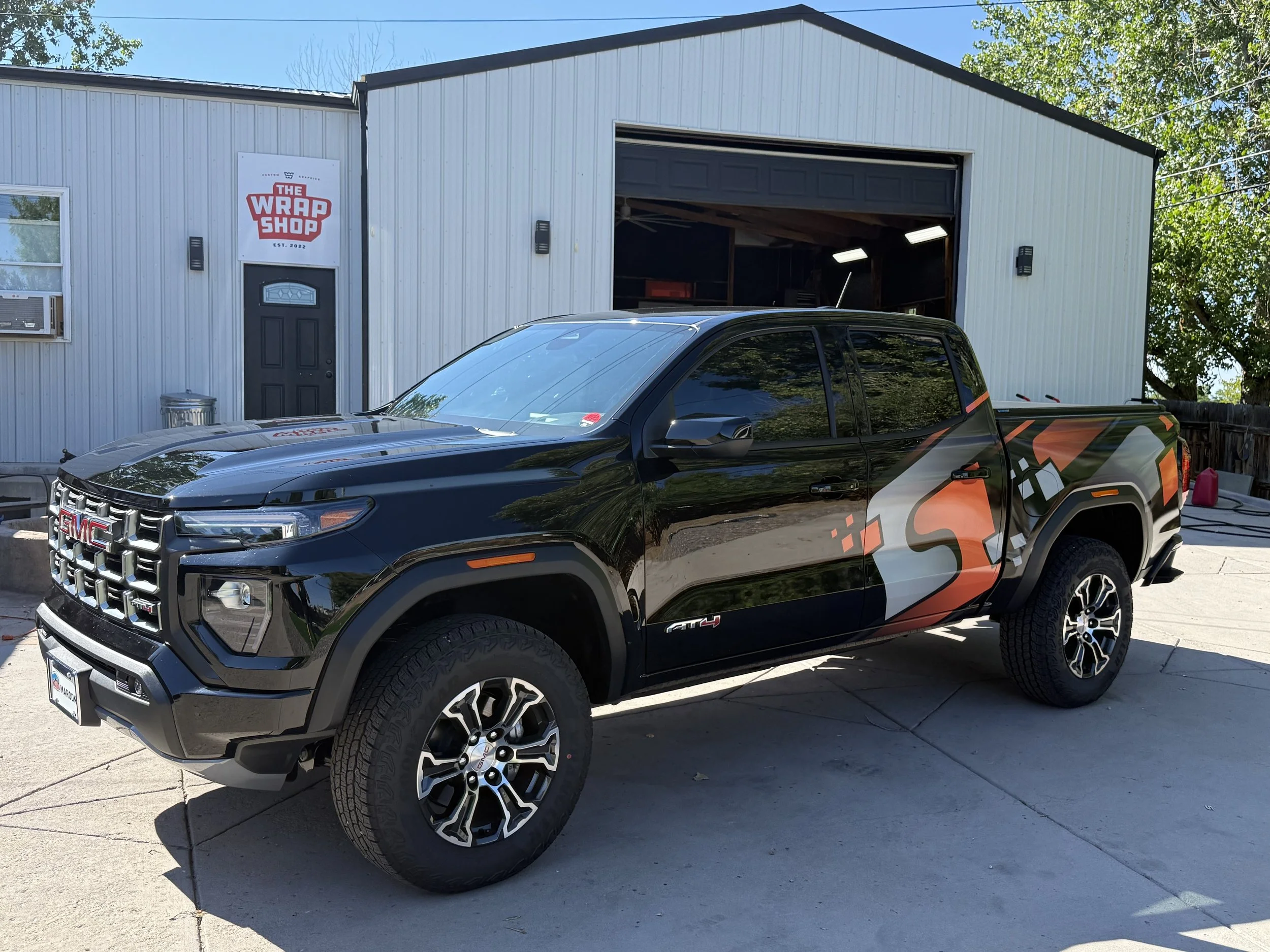 Black GMC pickup truck parked outside a building with grey metal siding. The building has a sign that reads 'The Wrap Shop'. The truck has a colorful graphic wrap on its side.