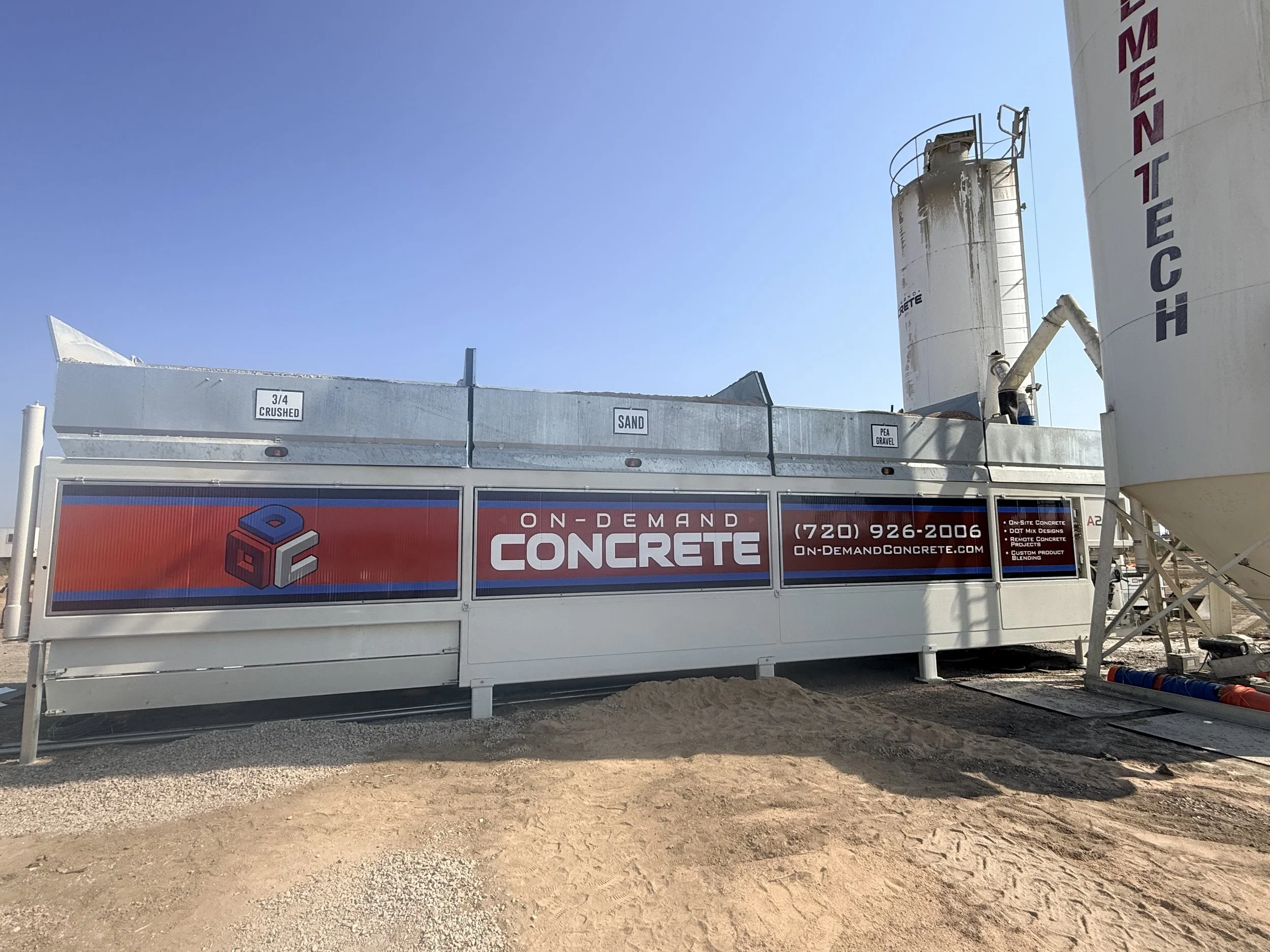 A portable concrete blend plant advertising on-demand concrete with contact information. Behind the blend plant are large cement silos with some labels, and the sky is clear and blue. The ground is sandy and gravelly.