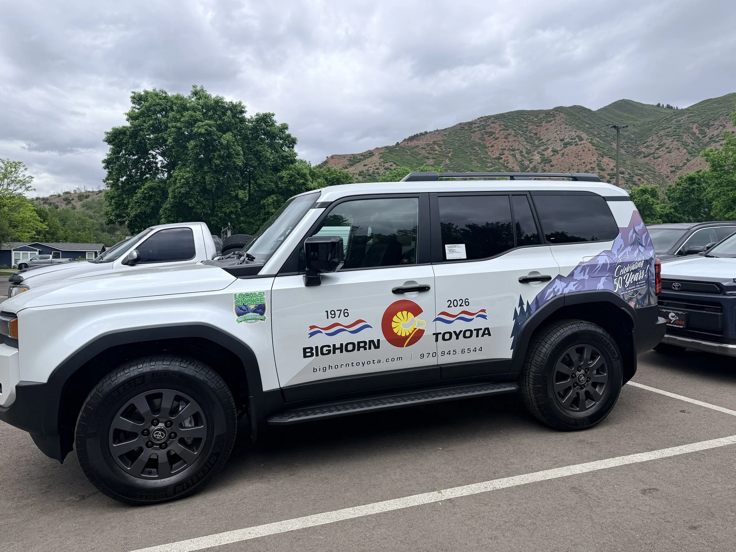 A white Toyota SUV parked in a parking lot with a mountainous landscape and cloudy sky in the background. The vehicle has promotional decals and text on its side, including logos and a phone number.
