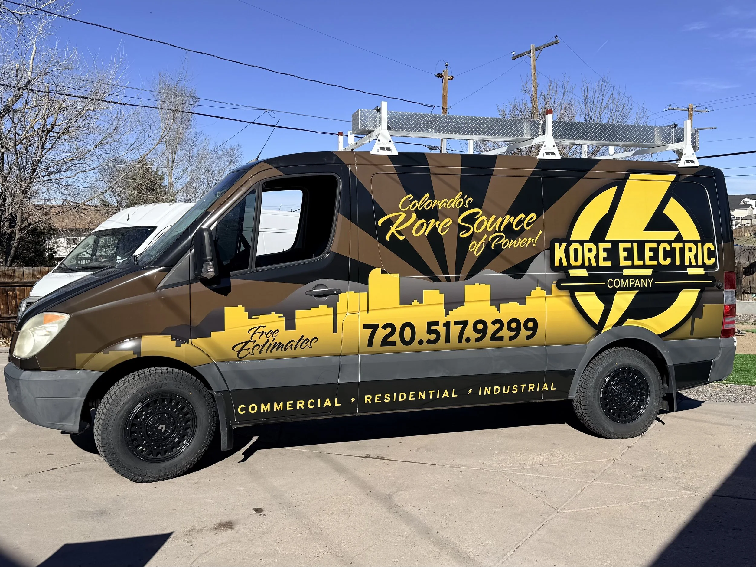 A company van for Kore Electric, displaying electrical services. The van has a brown, black, and yellow color scheme, and features a large logo with the text "Kore Electric" and a phone number. It is equipped with a metal ladder rack on top.