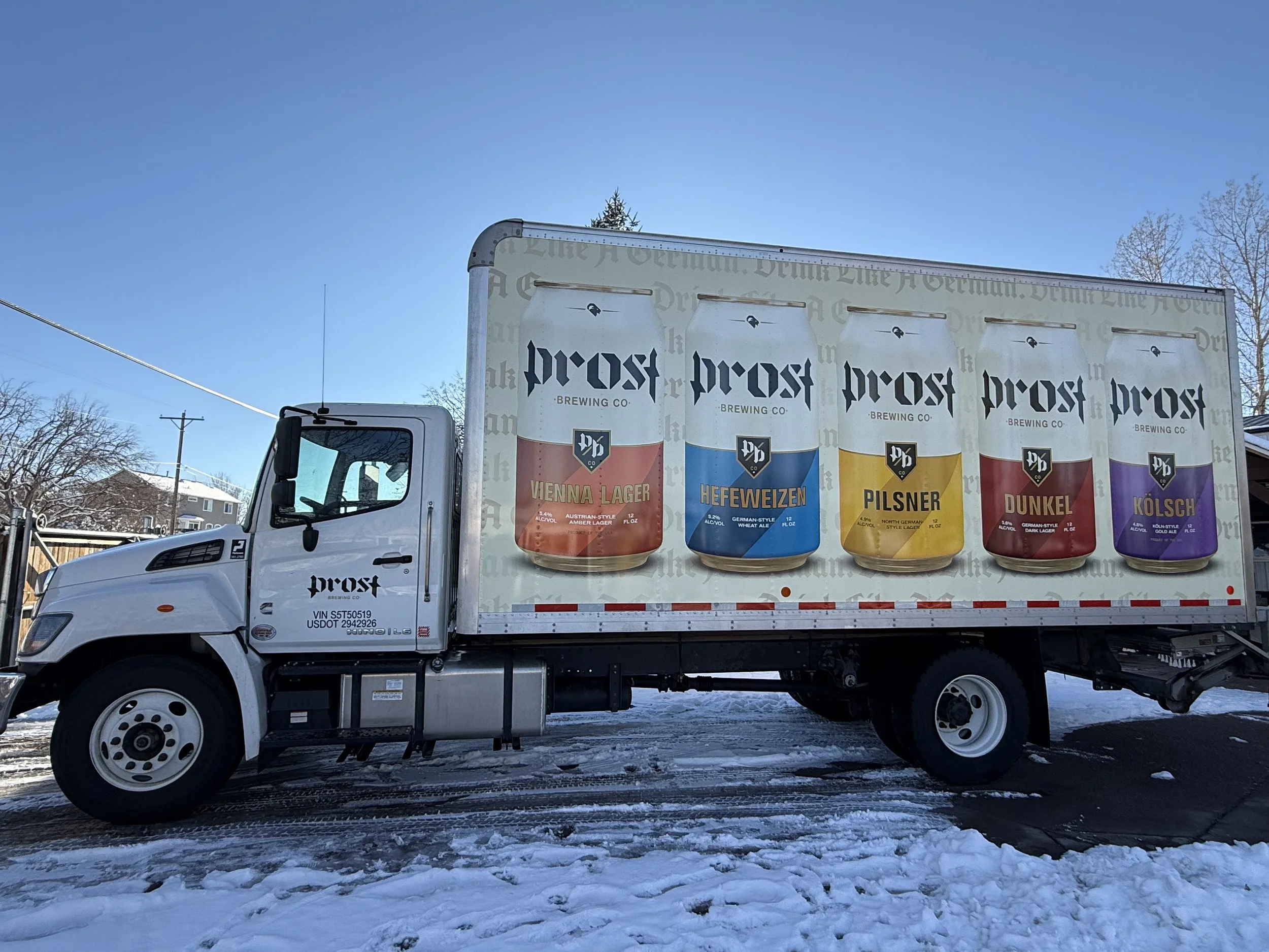 A white delivery truck with advertising for Prost Brewing Company, featuring images of five different beer cans on the side. The truck is parked on a snowy road with a clear blue sky and some trees in the background.