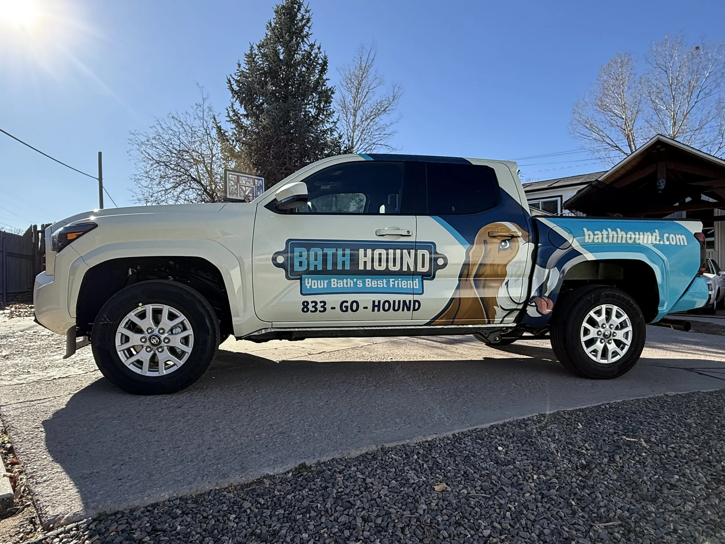A white pickup truck with Bath Hound advertisements parked on a gravel surface under a bright blue sky, with a basketball hoop, trees, and a house nearby.