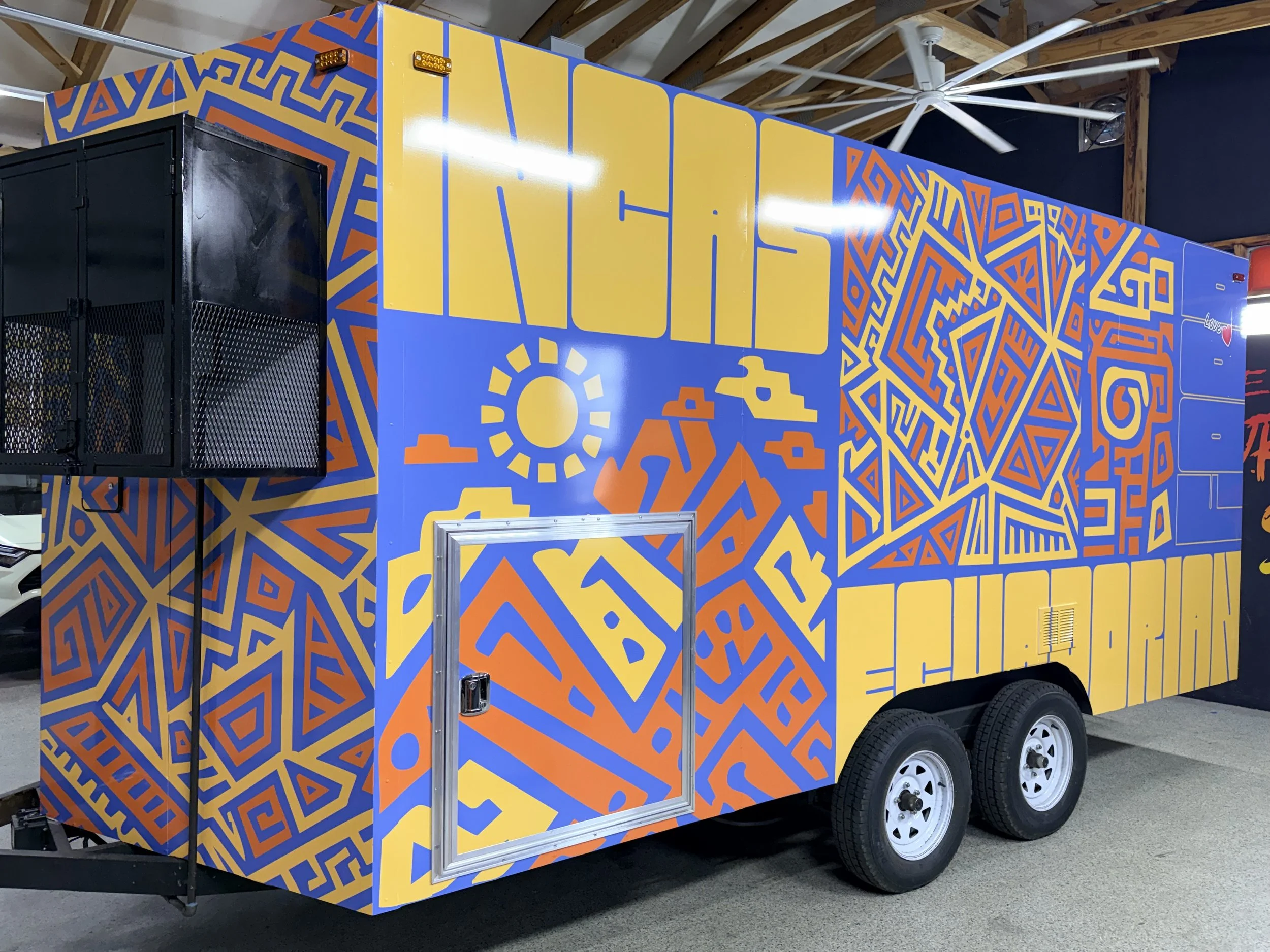 A trailer with a colorful geometric mural in blue, yellow, orange, and red, featuring various maze-like and abstract patterns. The trailer has a black enclosed section and is parked on a concrete surface inside a building with exposed wooden beams.