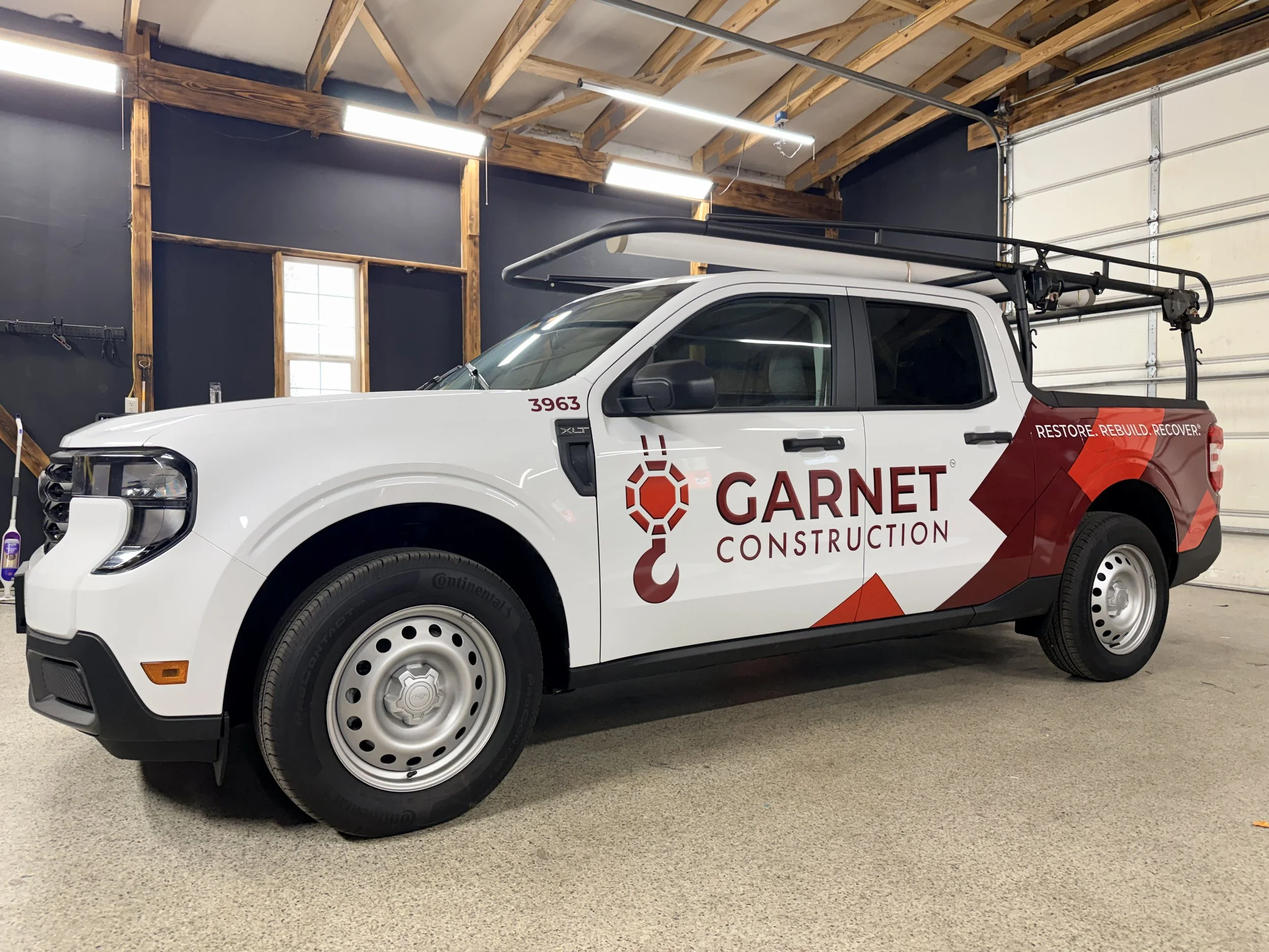 A white pickup truck with GARNET construction logo and slogan 'Restore. Rebuild. Recover.' parked inside a garage with wooden framing and black walls.