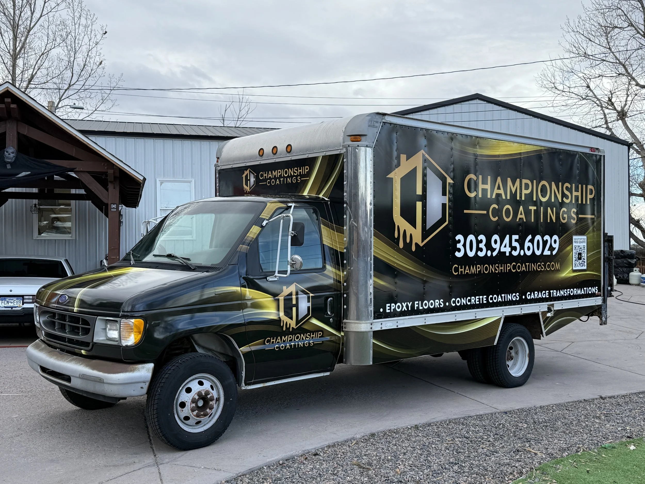 A black and yellow truck with branding for Championship Coatings, parked on a driveway in front of a light blue building with a metal roof. The truck advertises epoxy floors, concrete coatings, garage transformations, and has a phone number and websi