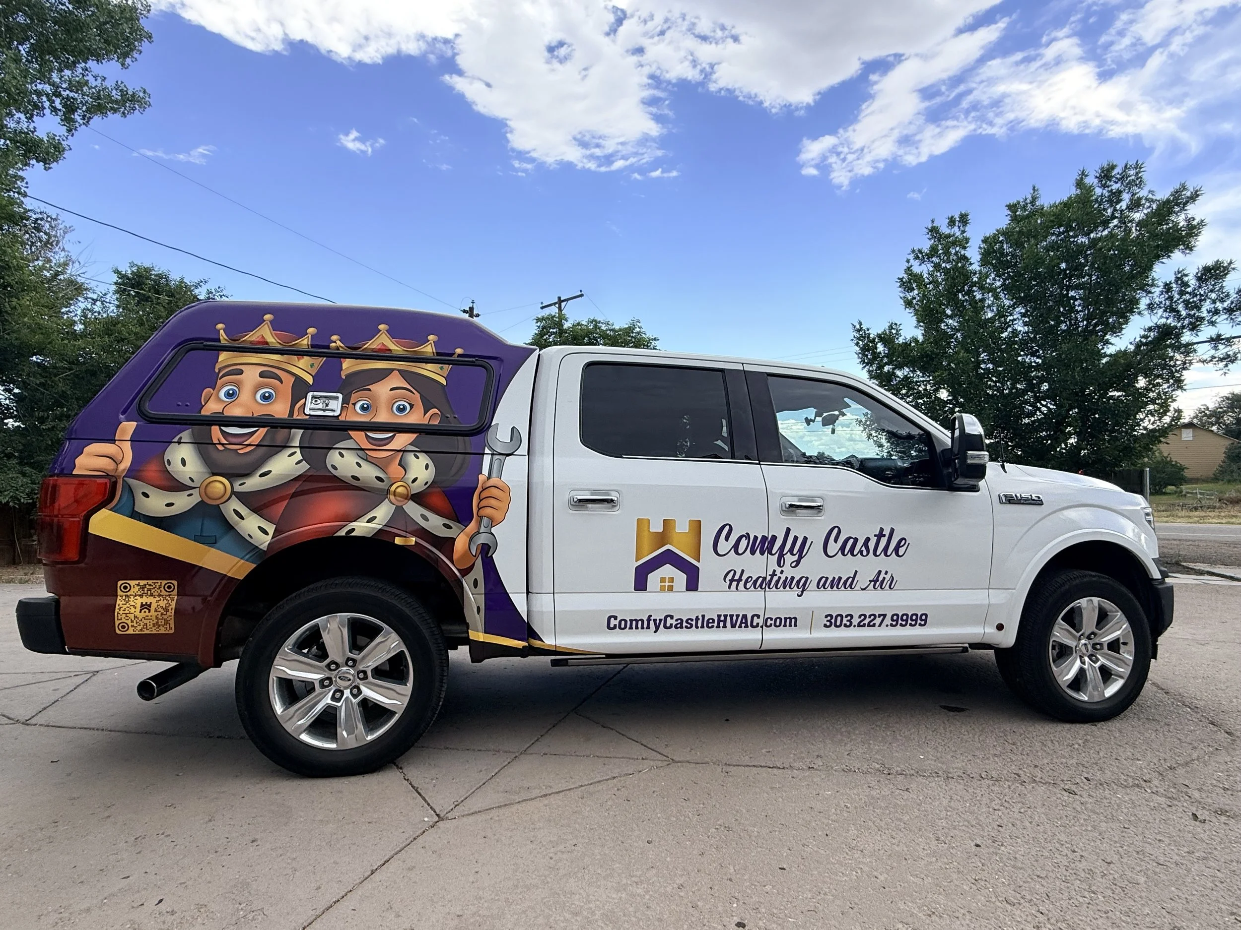A white work van with purple and yellow accents featuring cartoon characters of a king and queen on the side. The van has marketing text for Comfy Castle Heating and Air, including a website, phone number, and a QR code, parked on a concrete driveway