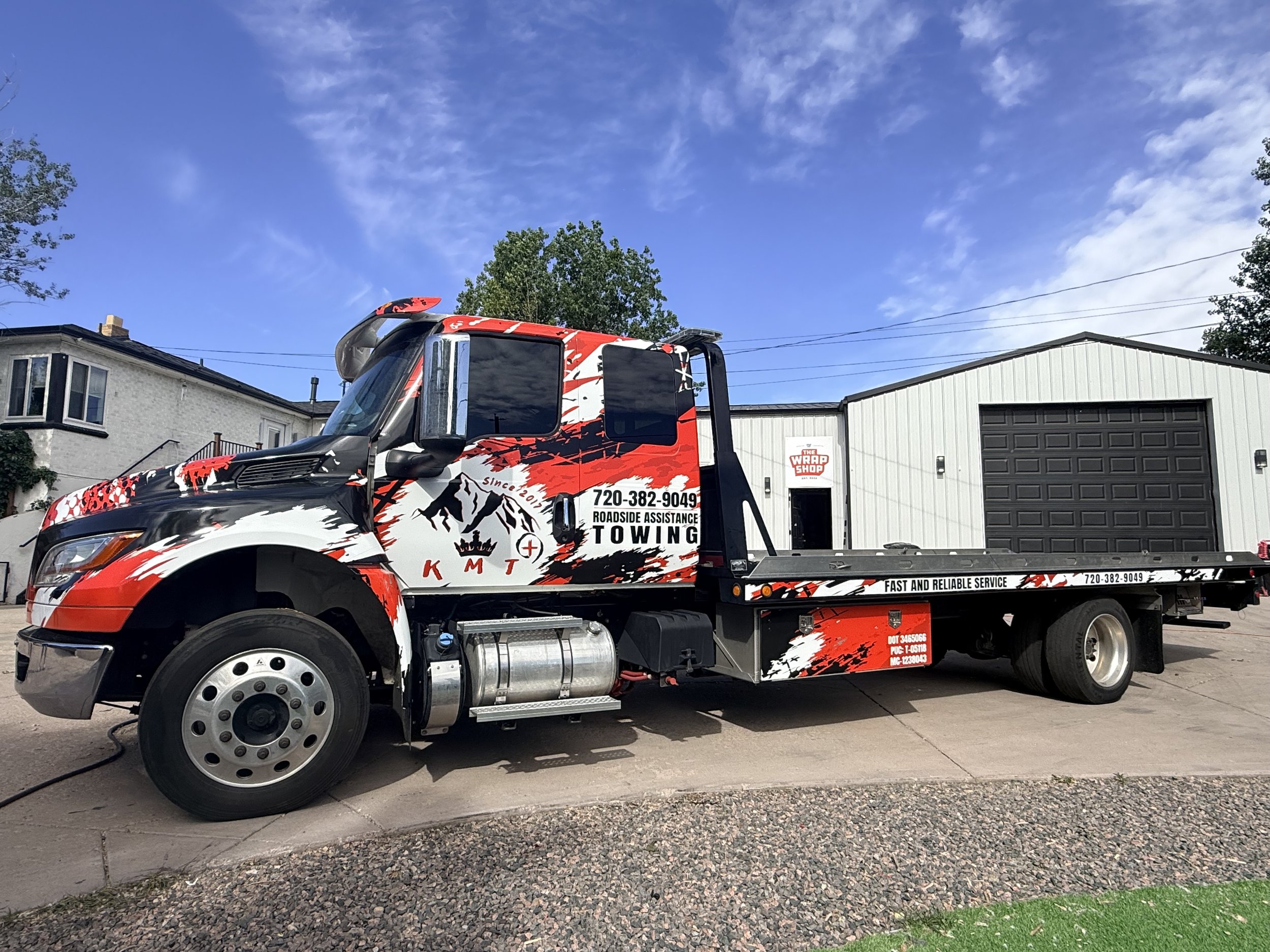 A tow truck parked on a driveway in front of a house and a garage, with a clear blue sky and some trees in the background.