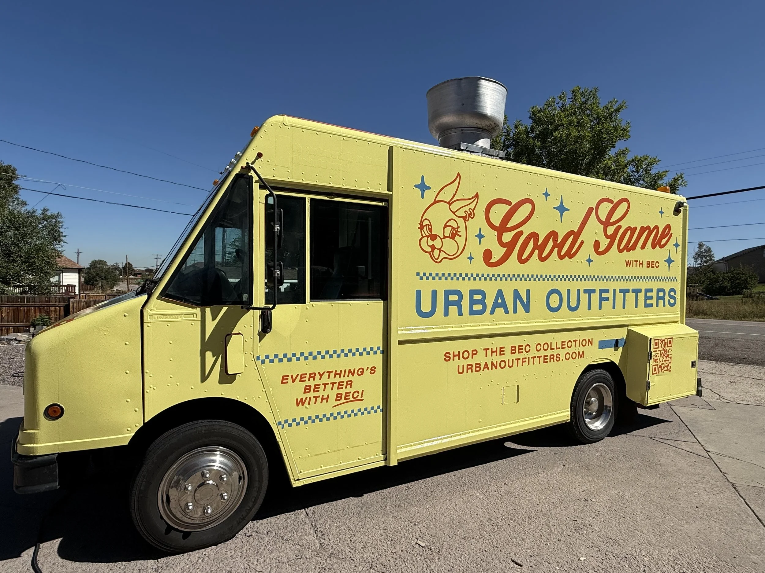 A yellow mobile food truck with a cartoon bunny logo and the words "Good Game" and "Urban Outfitters" on its side, parked outdoors against a clear blue sky with some trees and power lines in the background.