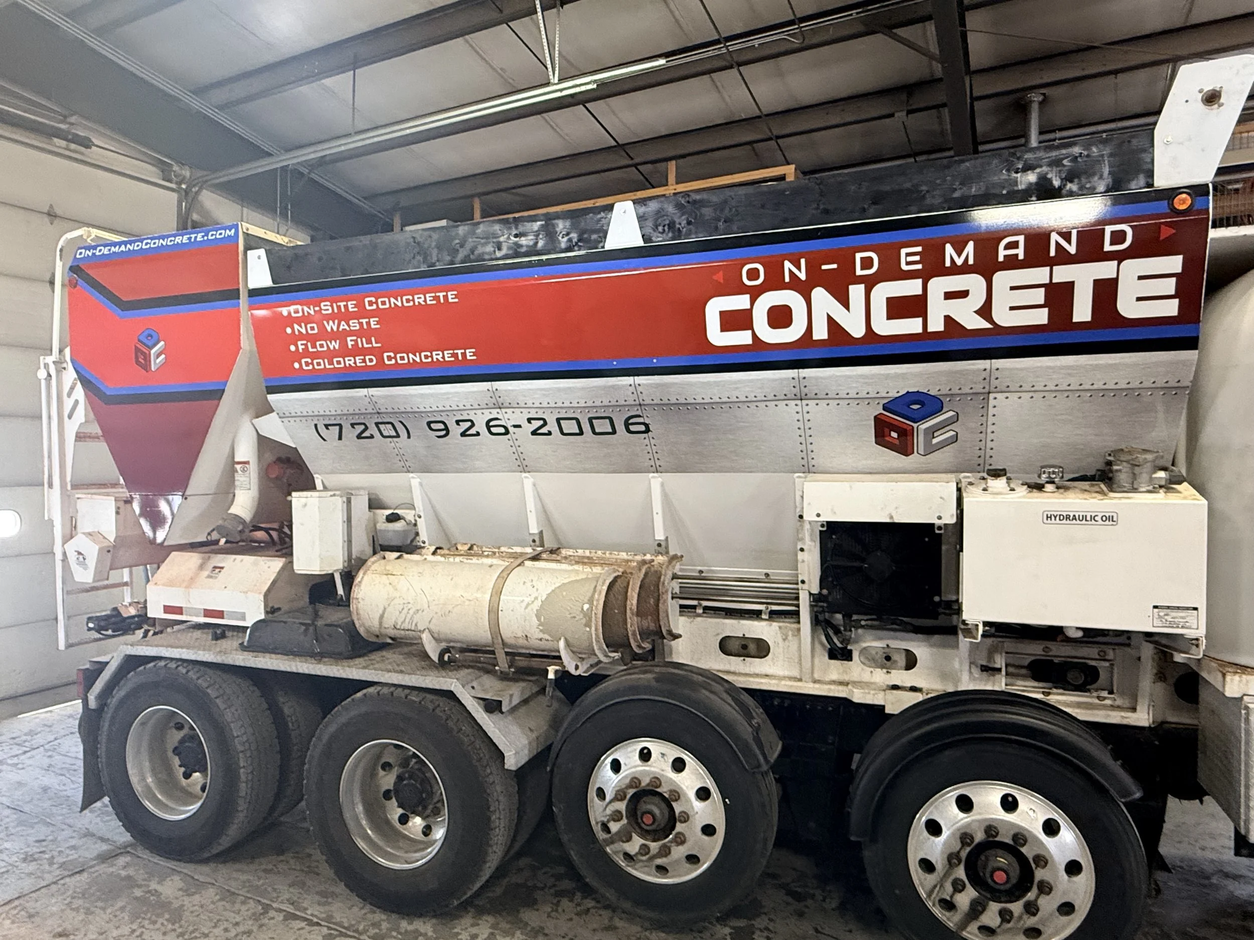 A large concrete truck with a sign reading "On-Demand Concrete" and instructions for on-site concrete, no waste, flow fill, and colored concrete. The truck has multiple wheels and a large aggregate drum, used for mixing and transporting concrete.