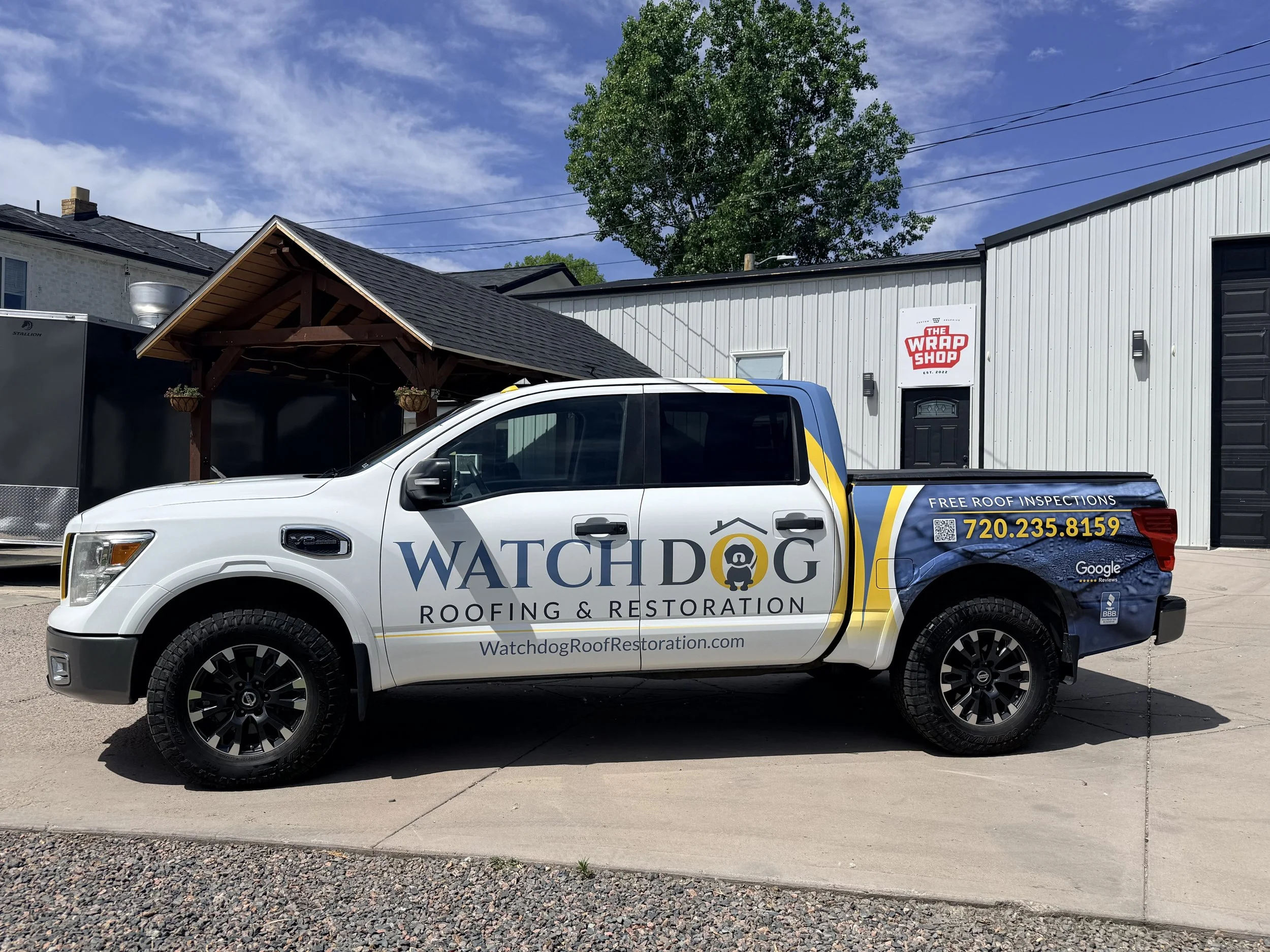 White pickup truck with Watchdog Roofing & Restoration branding parked in front of a commercial building with a sign that says The Wrap Shop. The building has a metal exterior and a sloped roof with a tree and blue sky in the background.