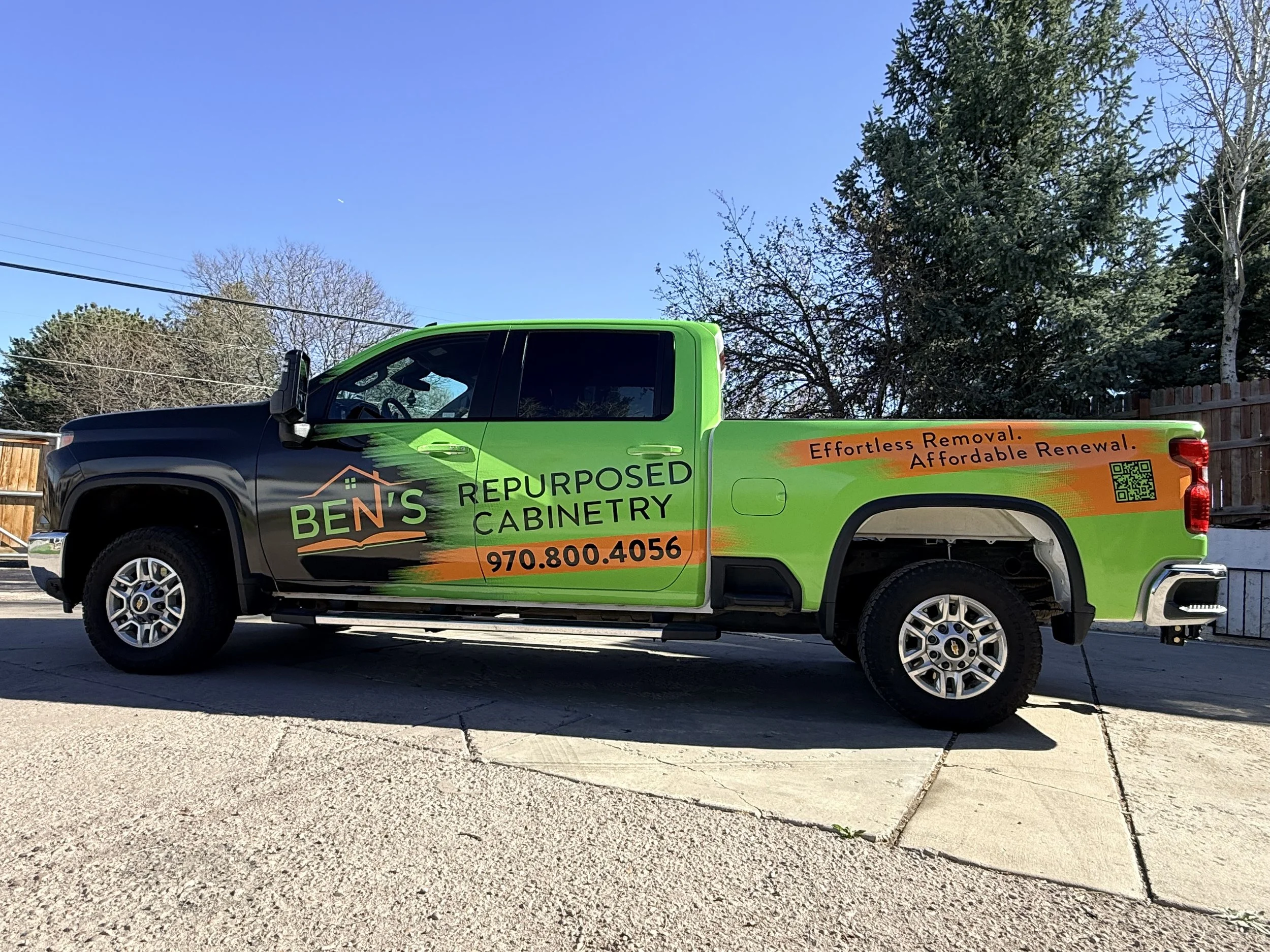 A green and black pickup truck with advertising on the side, parked on a concrete surface. The truck promotes a cabinetry business called Ben's Repurposed Cabinetry, with a slogan 'Effortless Removal. Affordable Renewal.' and a QR code. In the backgr
