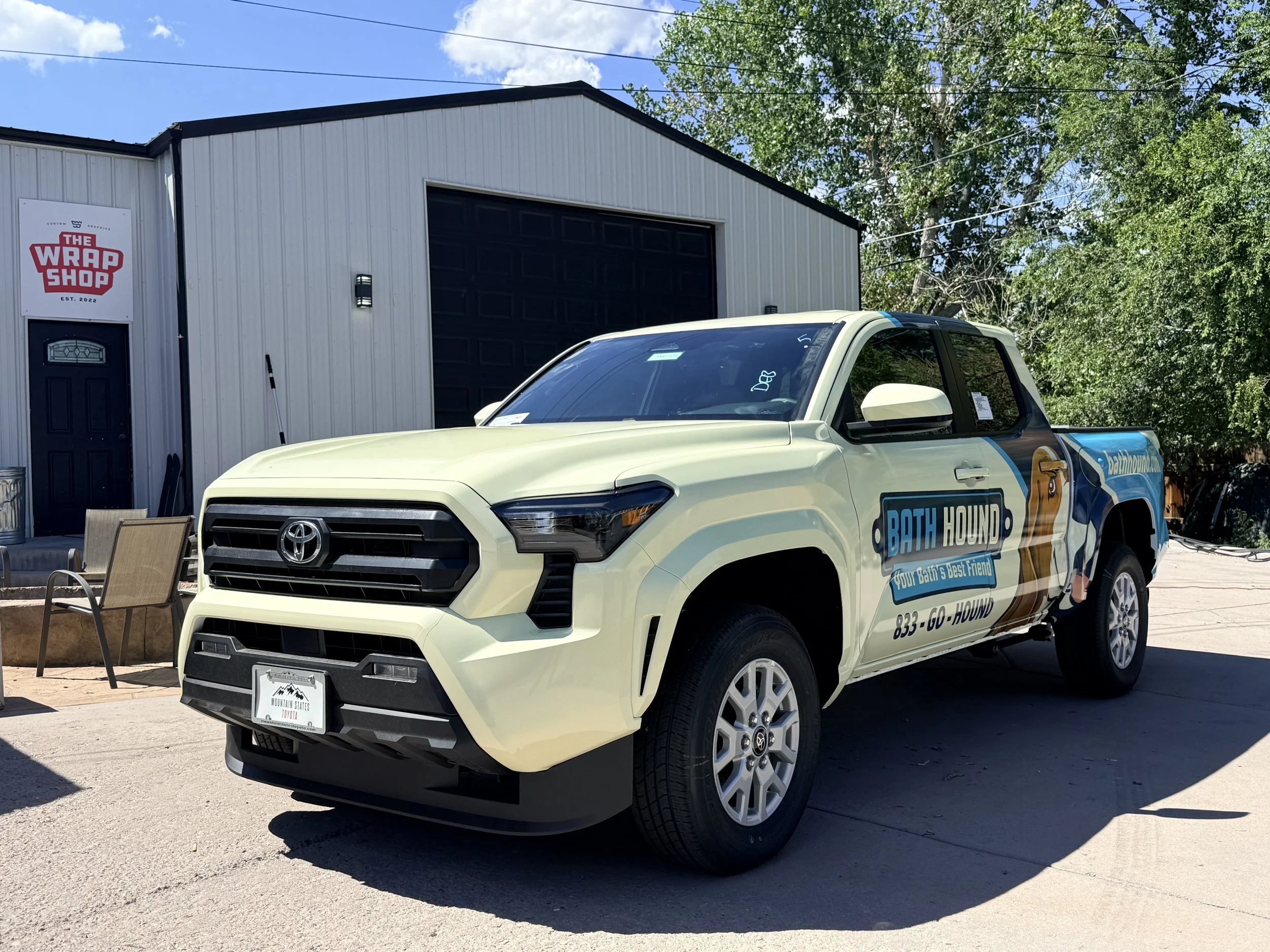 Front view of a white Toyota pickup truck with branding for Bath Hound, parked in front of a metal building with a sign for The Wrap Shop, established in 2022. The building has a black garage door and a black door, with trees and power lines in the b