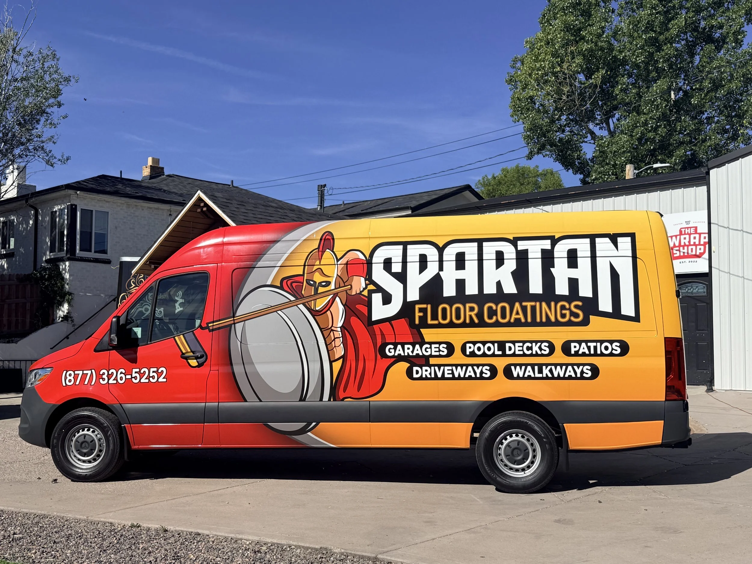 A red delivery van with advertising for Skarpton Floor Coatings, featuring a cartoon warrior with a helmet and shield, parked on a concrete street in a residential neighborhood with houses, trees, and a clear blue sky.