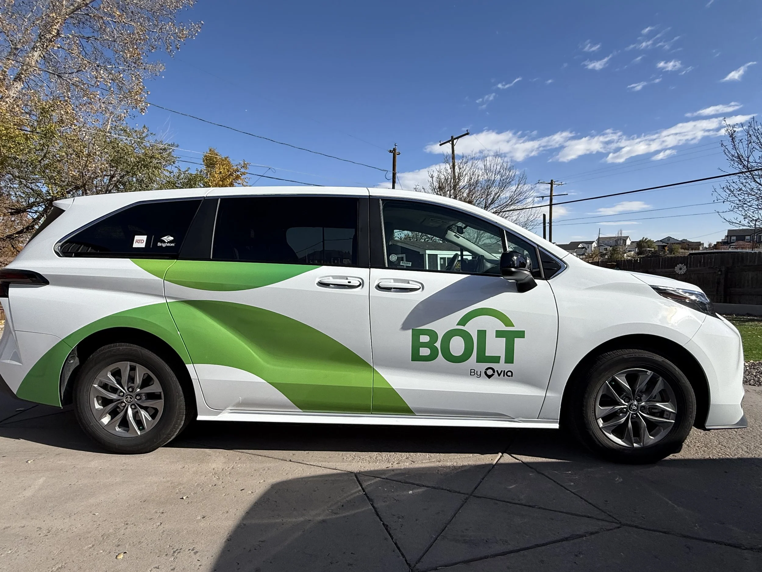 A white and green Bolt electric vehicle parked on a residential street under a blue sky with some clouds and power lines. Trees and houses are visible in the background.