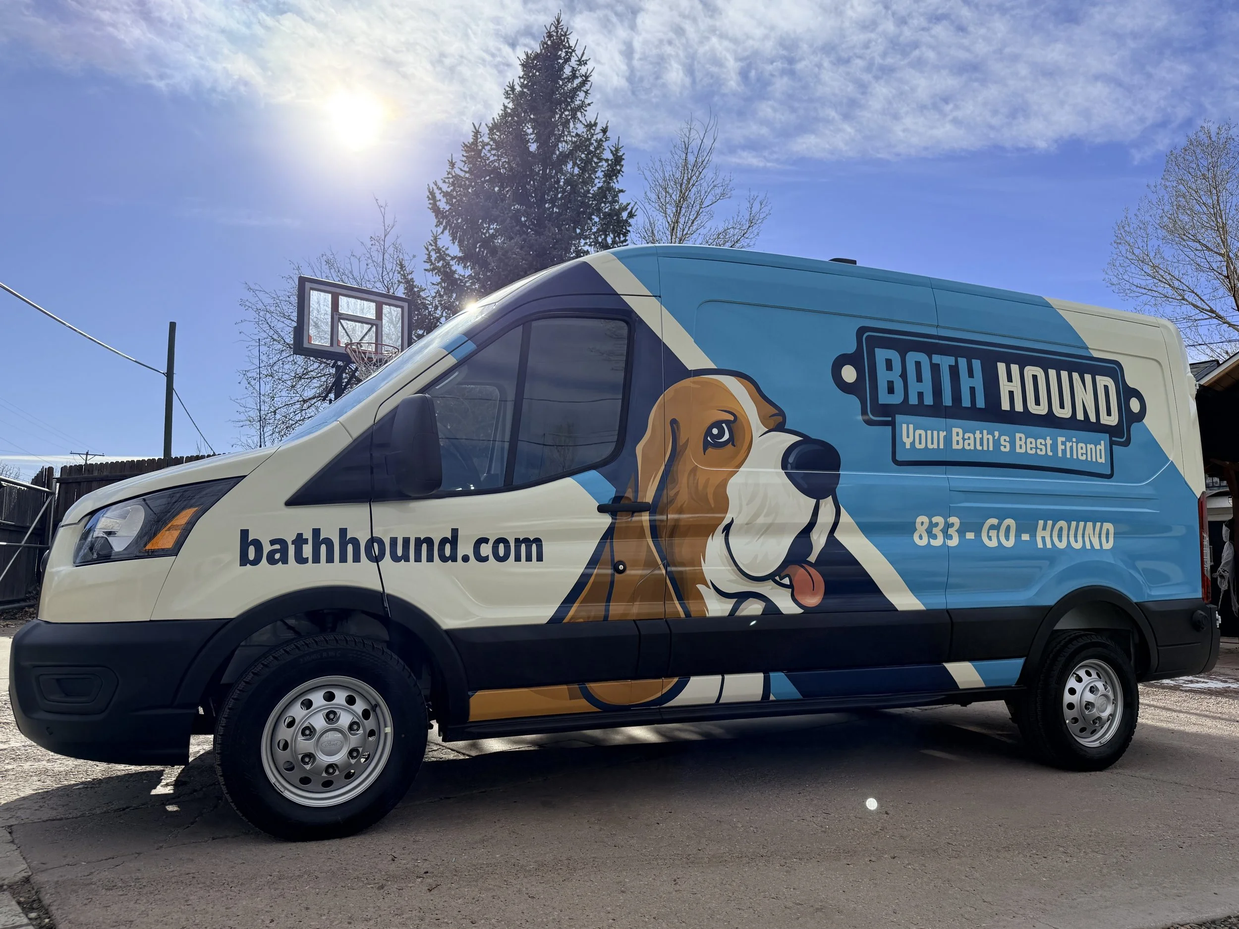 A Bath Hound service van parked outside with a basketball hoop and goalpost in the background on a sunny day.