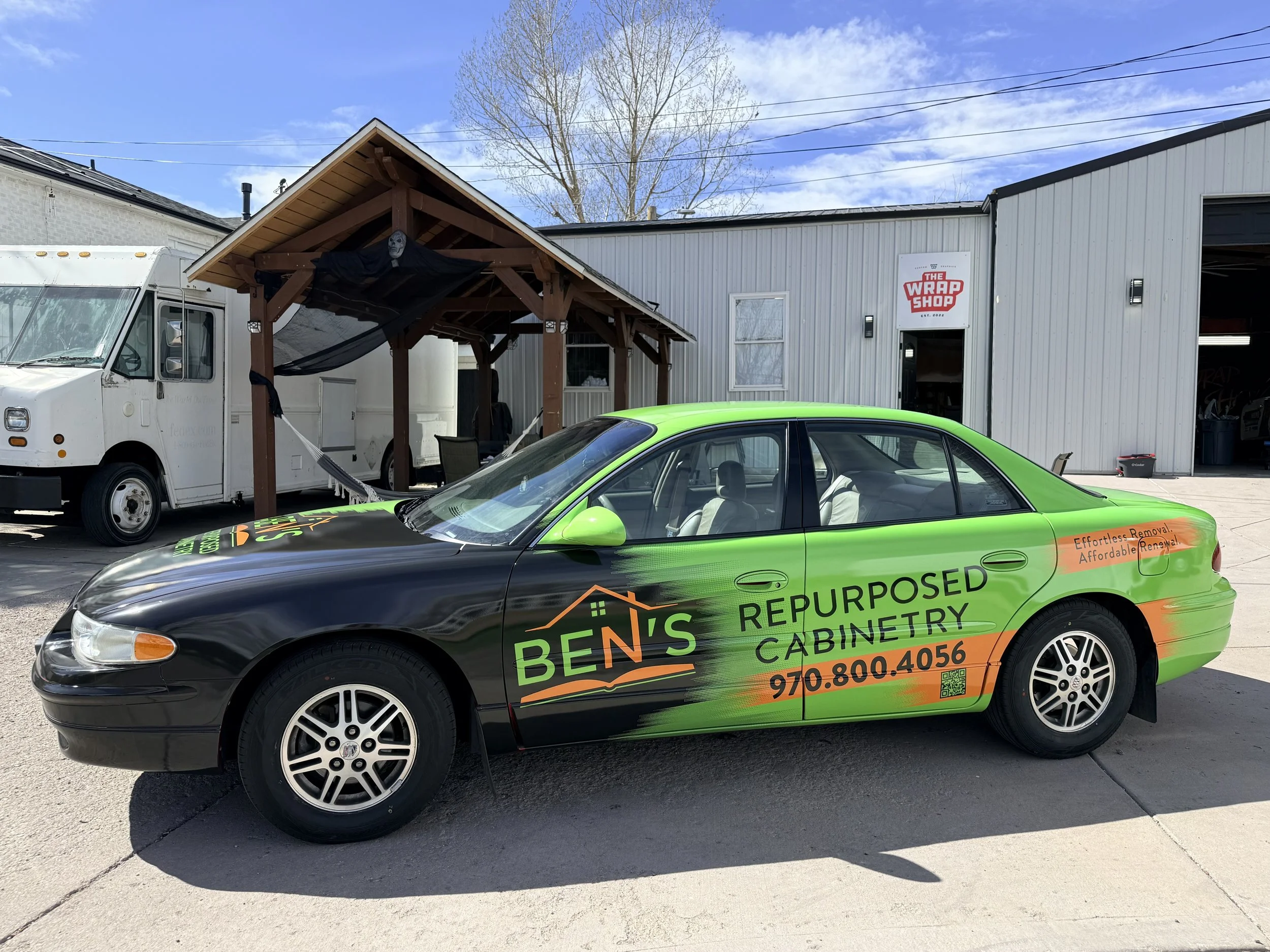 A green and black car with the company name 'Ben's RePurpose Cabinetry' and contact information parked in front of a building with a sign that reads 'The Wrap Shop'. There is a white food truck and a wooden structure with a skull decoration and a bla
