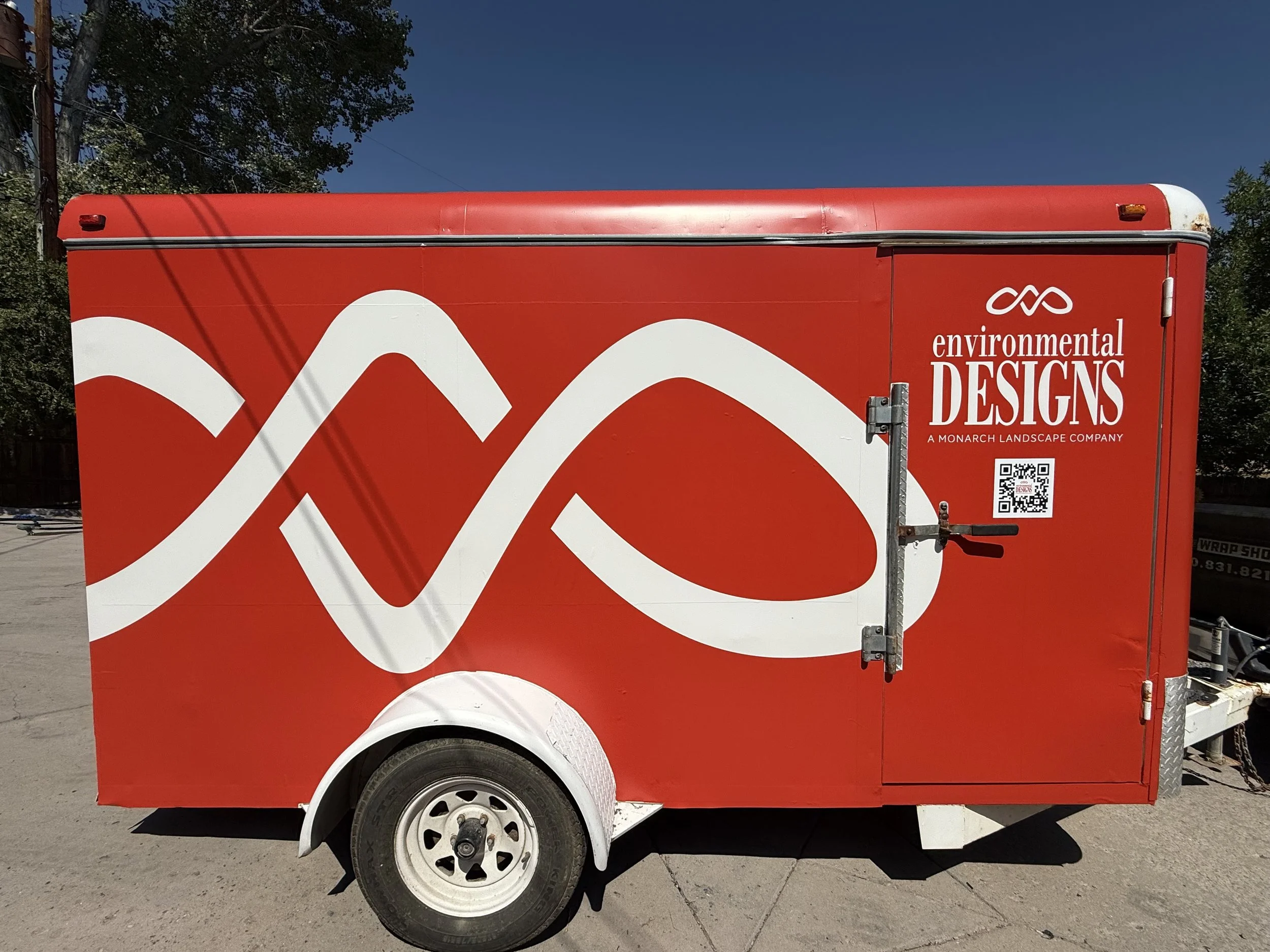 Red mobile food trailer with white decorative wave-like patterns and white text that reads 'Environmental Designs, a Monarch Landscape Company'. The trailer has a QR code, a small vent, a door latch, and is parked outdoors on a paved surface with tre