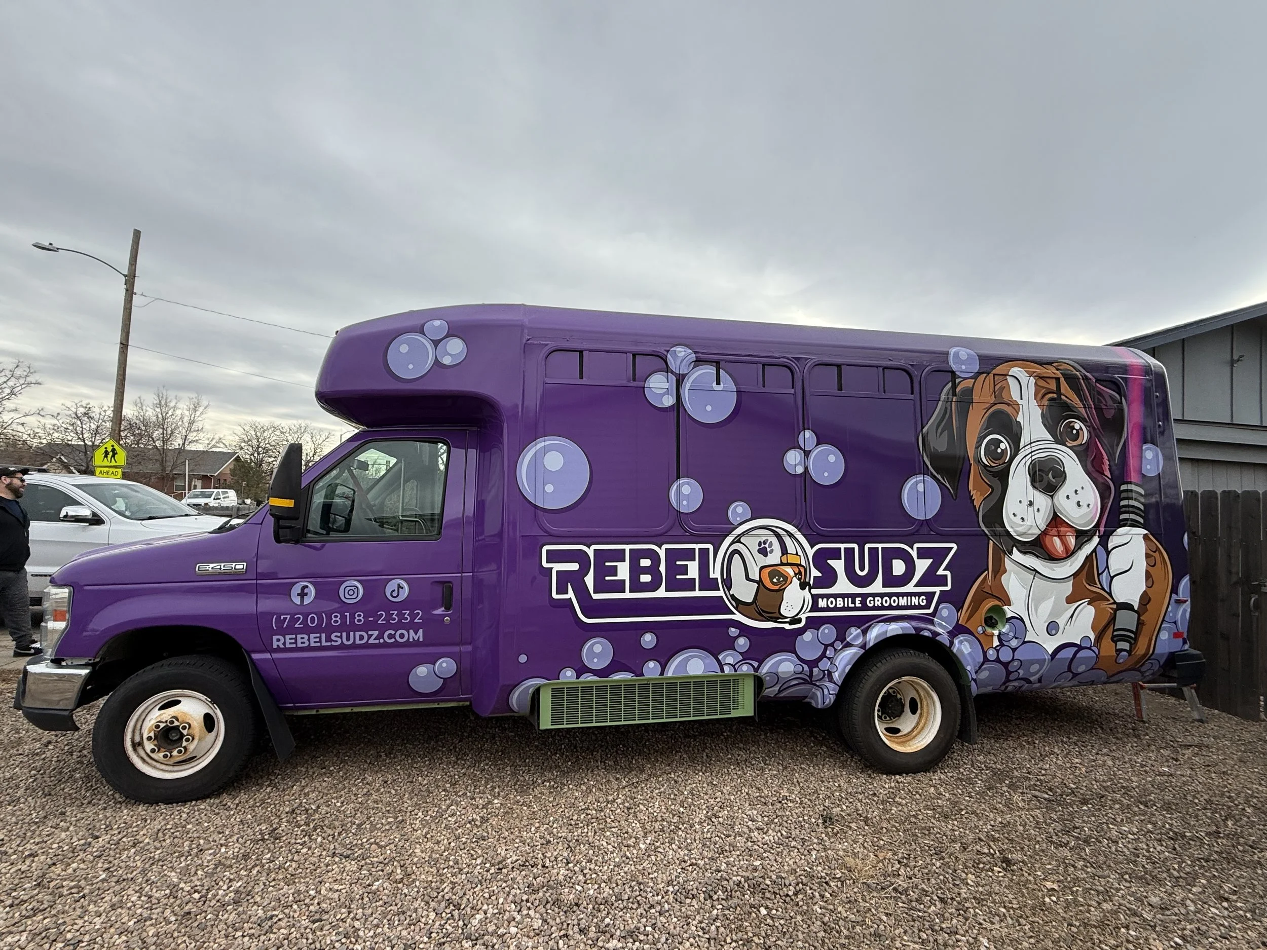 A purple mobile grooming van with cartoon dog graphics and bubbles, parked in a lot with a man standing nearby.