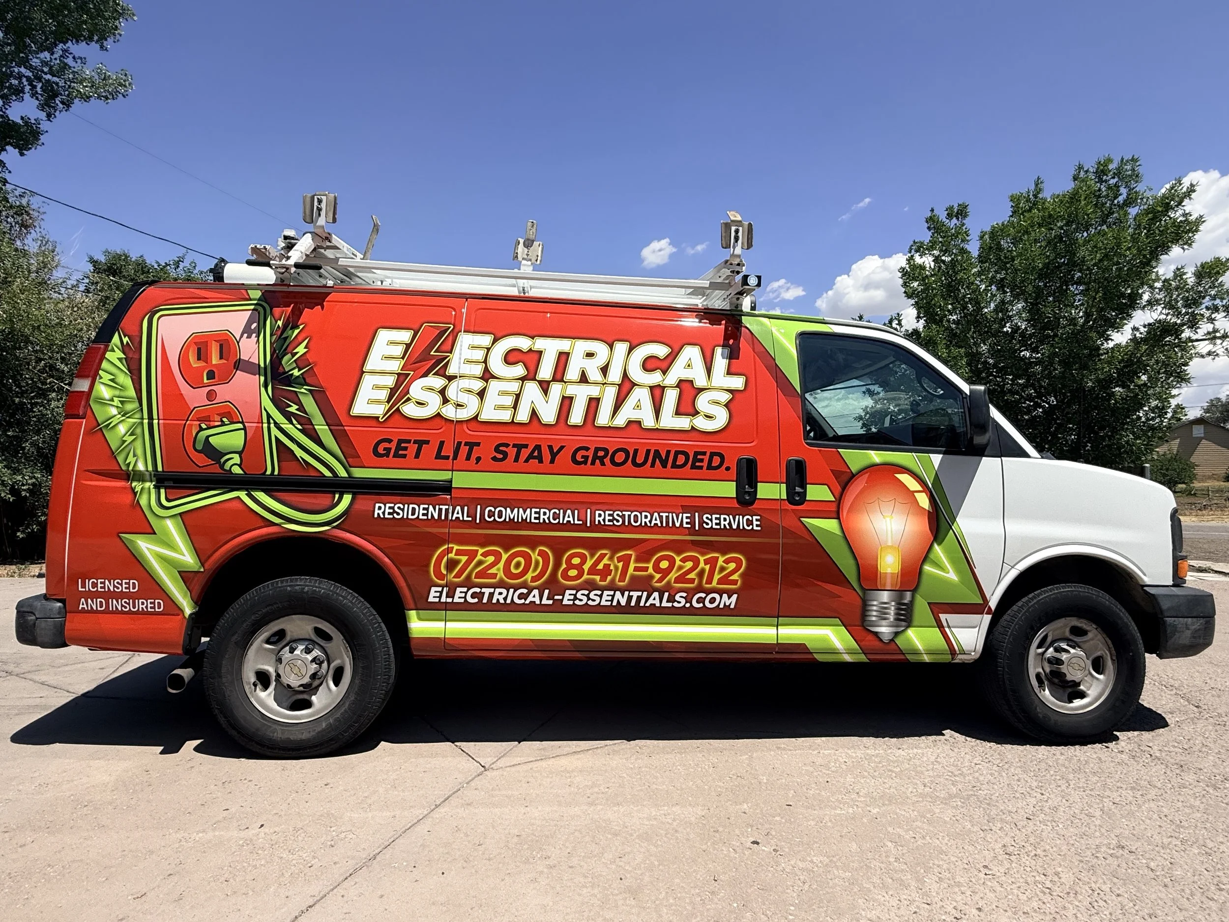 Red and green electrical service van with company branding and contact information parked on a driveway under a clear blue sky, surrounded by trees.