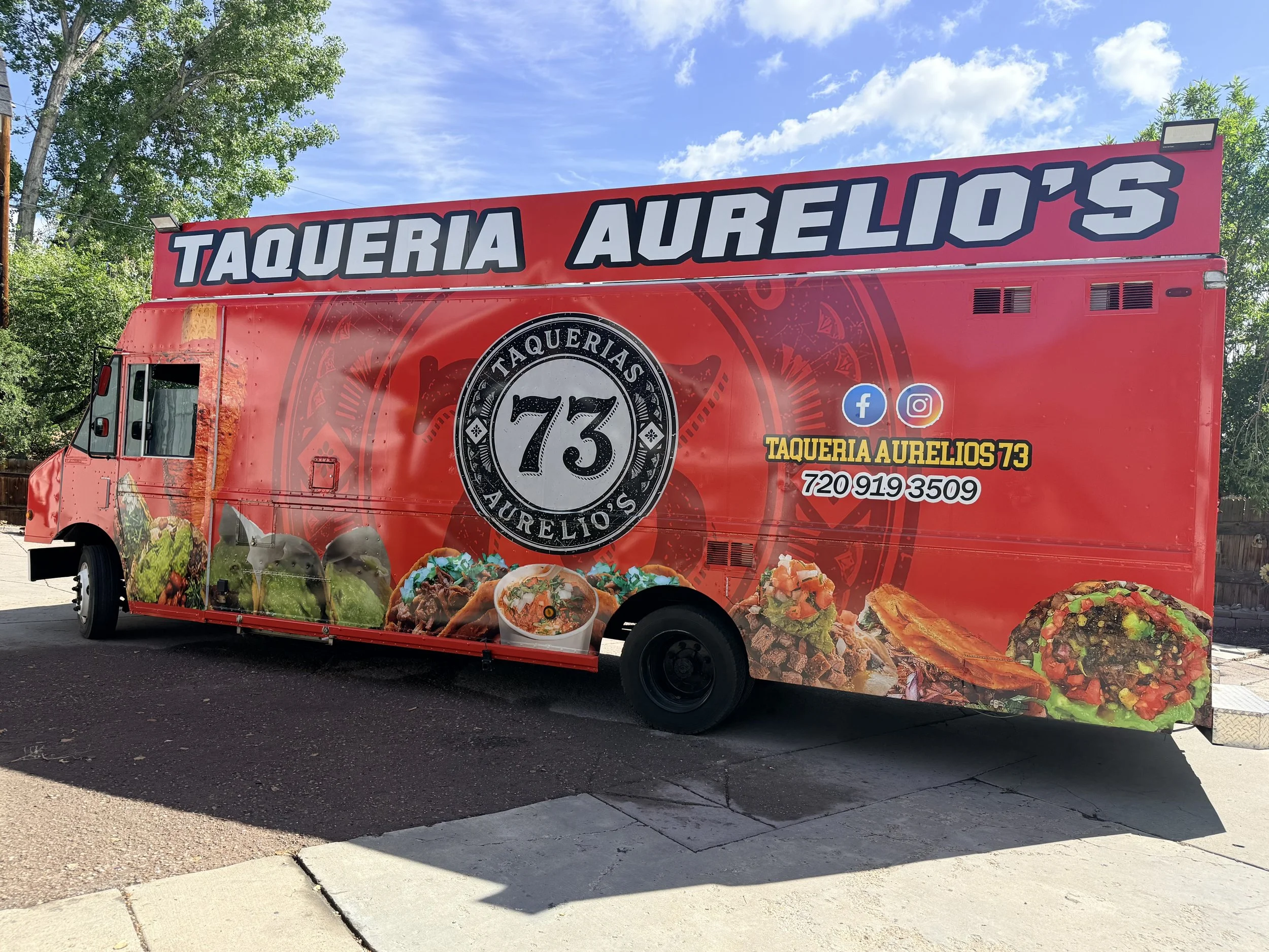 A red food truck with a sign that reads 'Taqueria Aurelio's' and branding for Facebook and Instagram. The truck features images of Mexican food such as tacos and nachos, parked outdoors under a blue sky with clouds and trees.